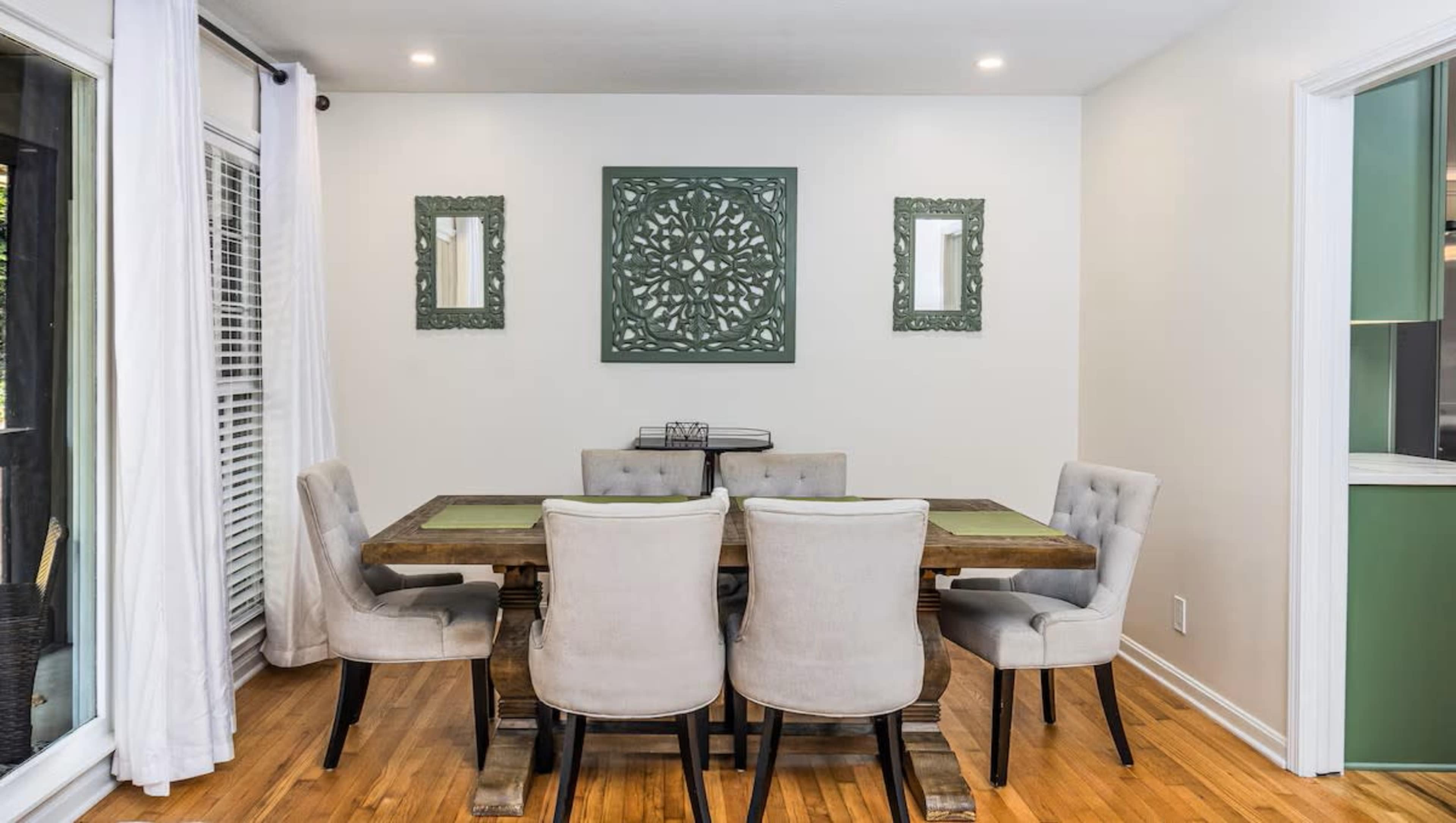 A dining room features a wooden table surrounded by six upholstered chairs, with decorative wall frames and a large piece of art on the wall.