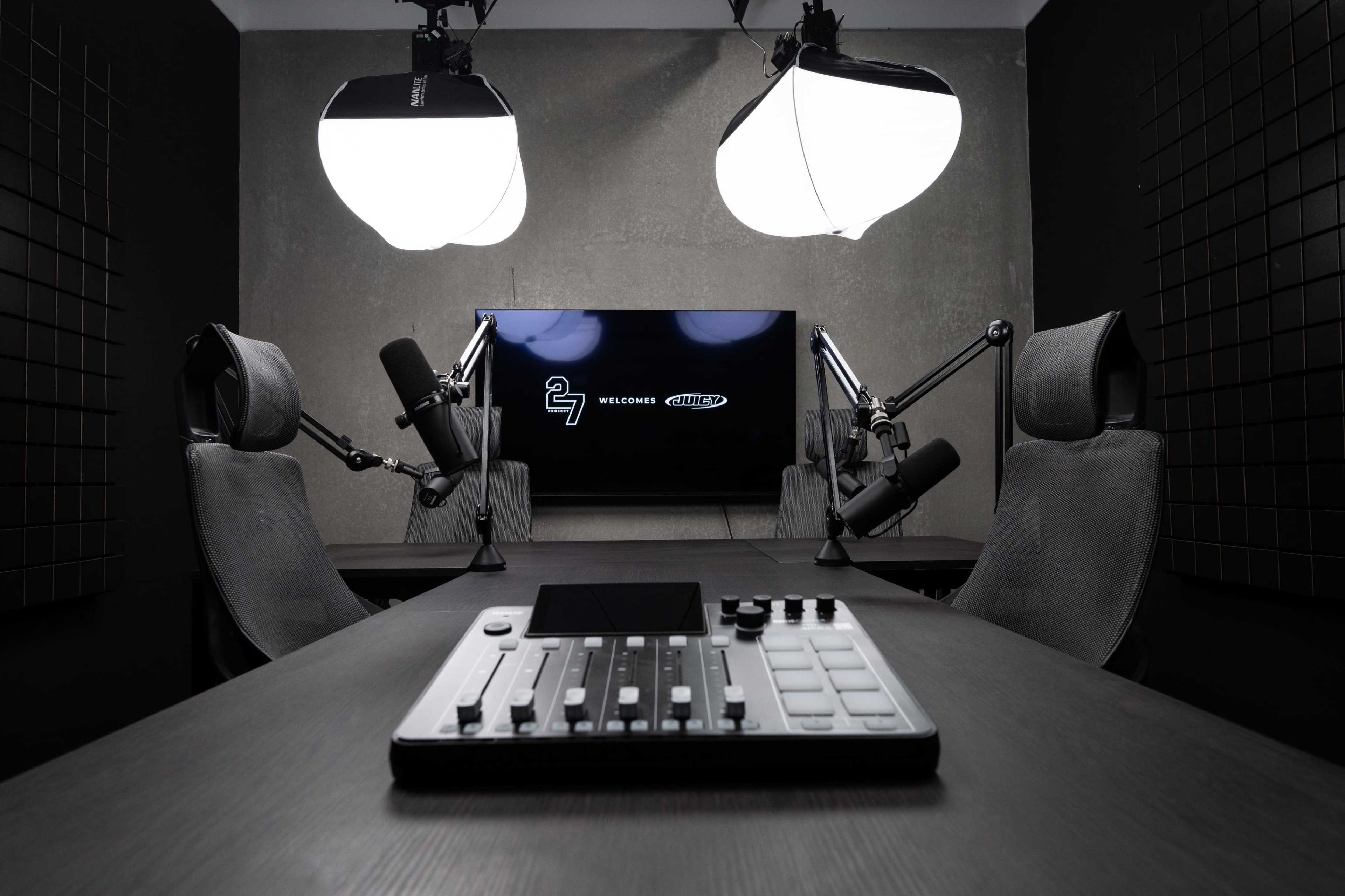 The image shows a podcasting studio equipped with two chairs, microphones on adjustable arms, a control panel, and a large screen displaying a welcome message.