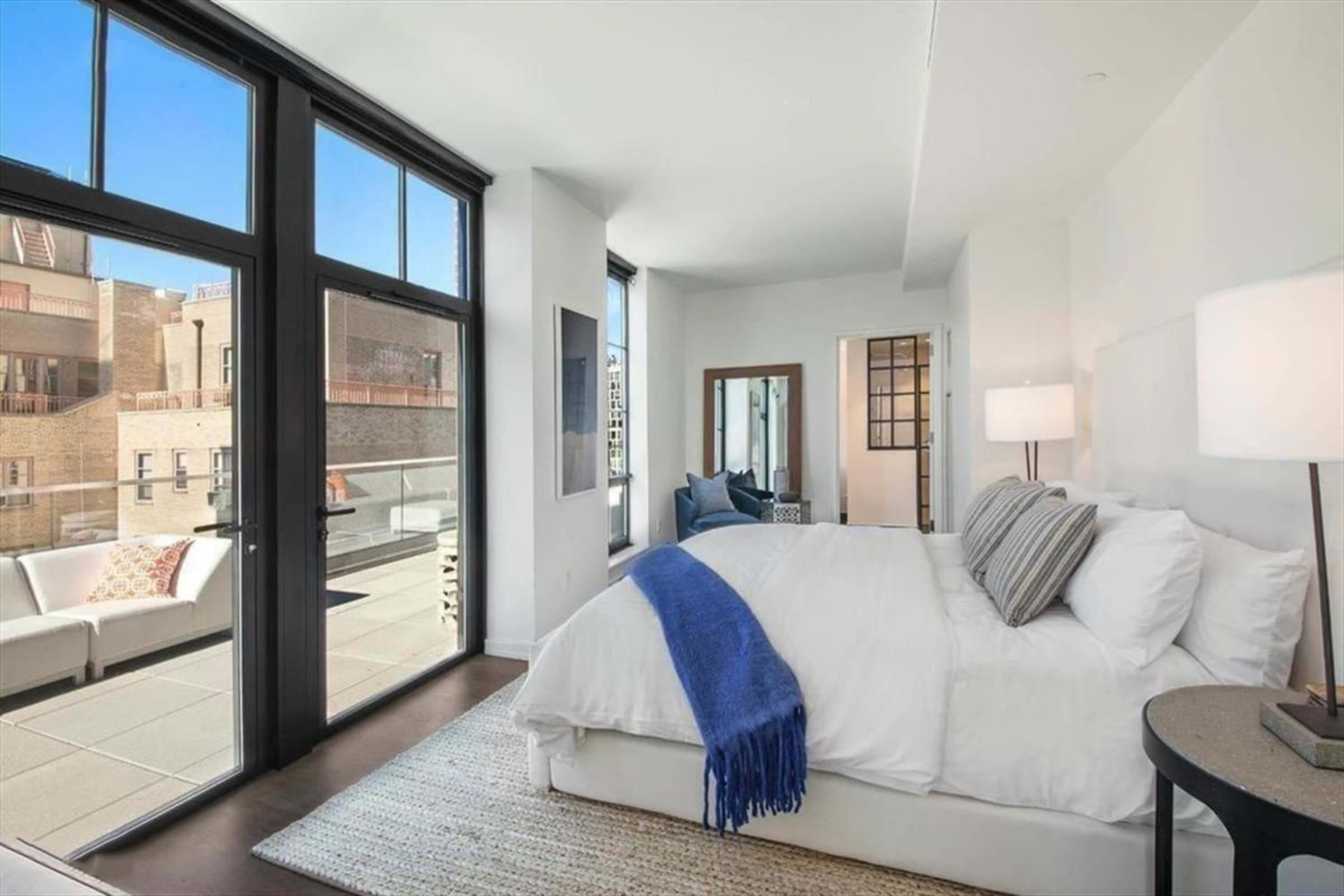 A modern bedroom features a large bed, a view of an outdoor balcony, and ample natural light from floor-to-ceiling windows.