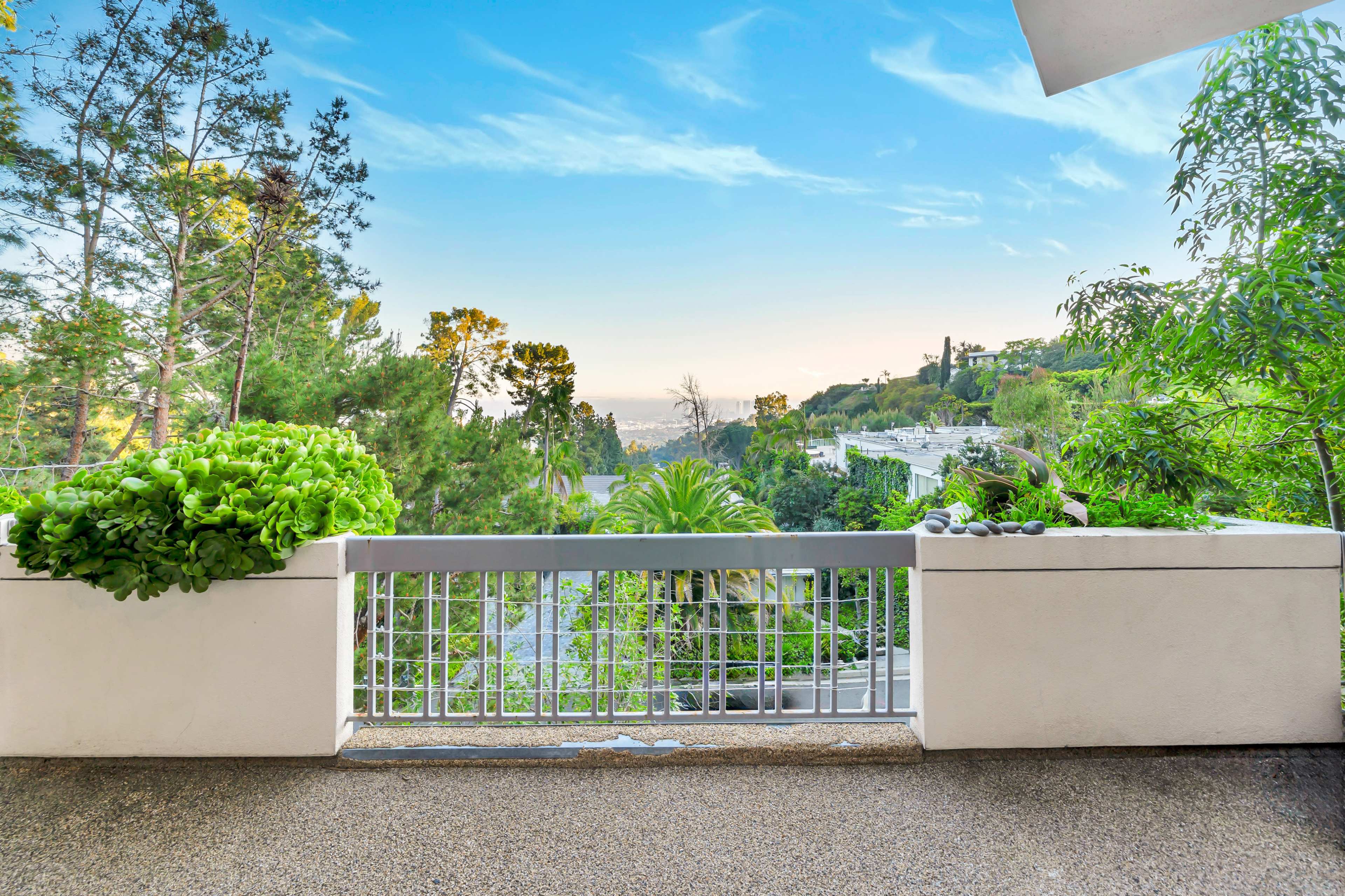 A balcony railing overlooks a lush green landscape with trees and distant hills under a clear sky.