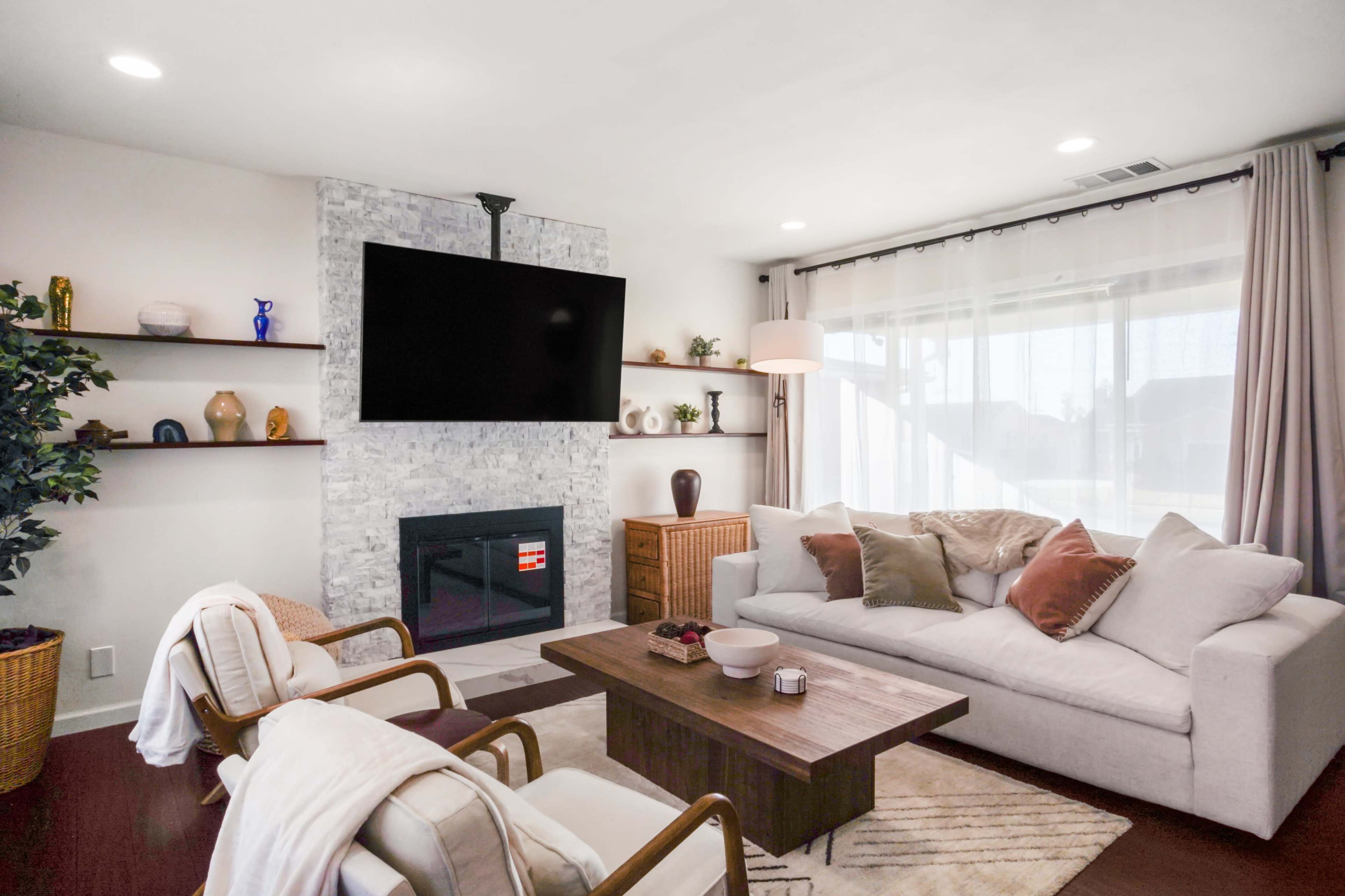 A modern living room with a white sofa, a wooden coffee table, two armchairs, a stone fireplace, and shelves displaying various decorative items.