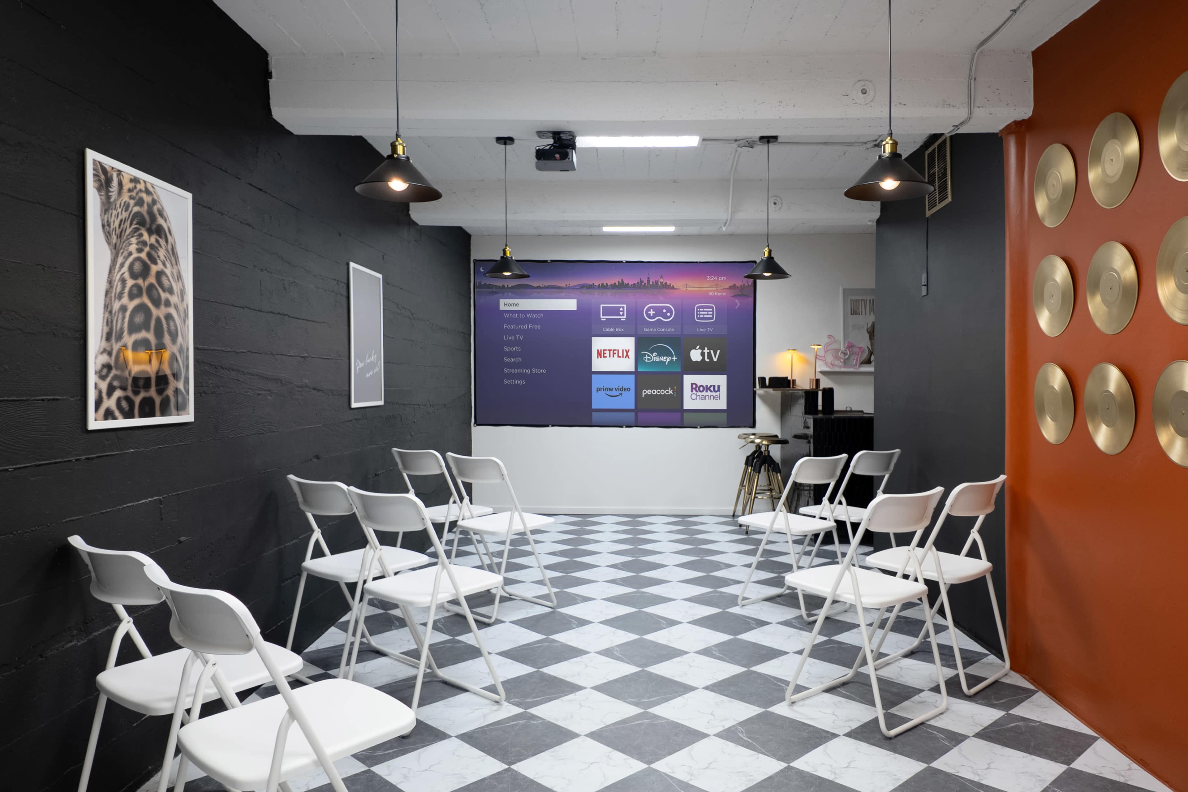 The image shows a modern room with a checkered floor, a wall featuring a screen displaying various streaming services, and rows of white folding chairs arranged for an event.