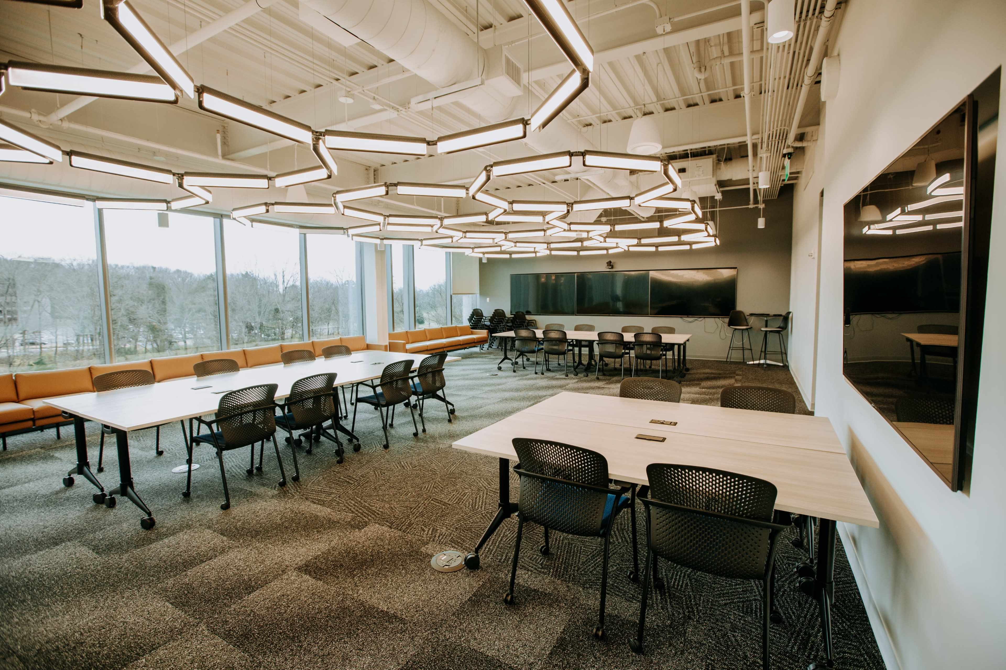 The image shows a modern conference room with multiple tables arranged for collaboration, large windows letting in natural light, and sleek lighting fixtures on the ceiling.