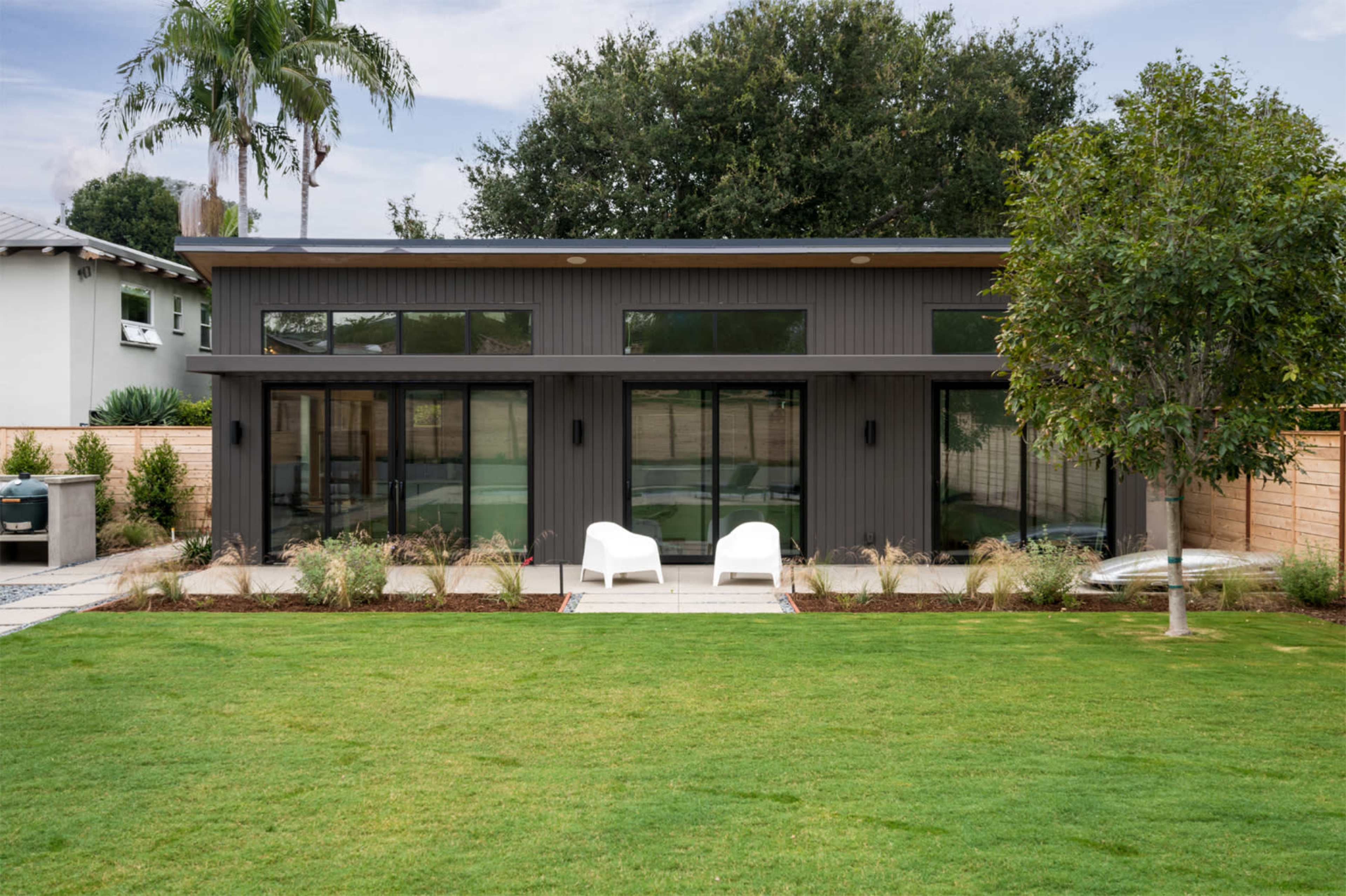 A modern house with large glass windows is surrounded by a lawn and features two white chairs on a stone patio.