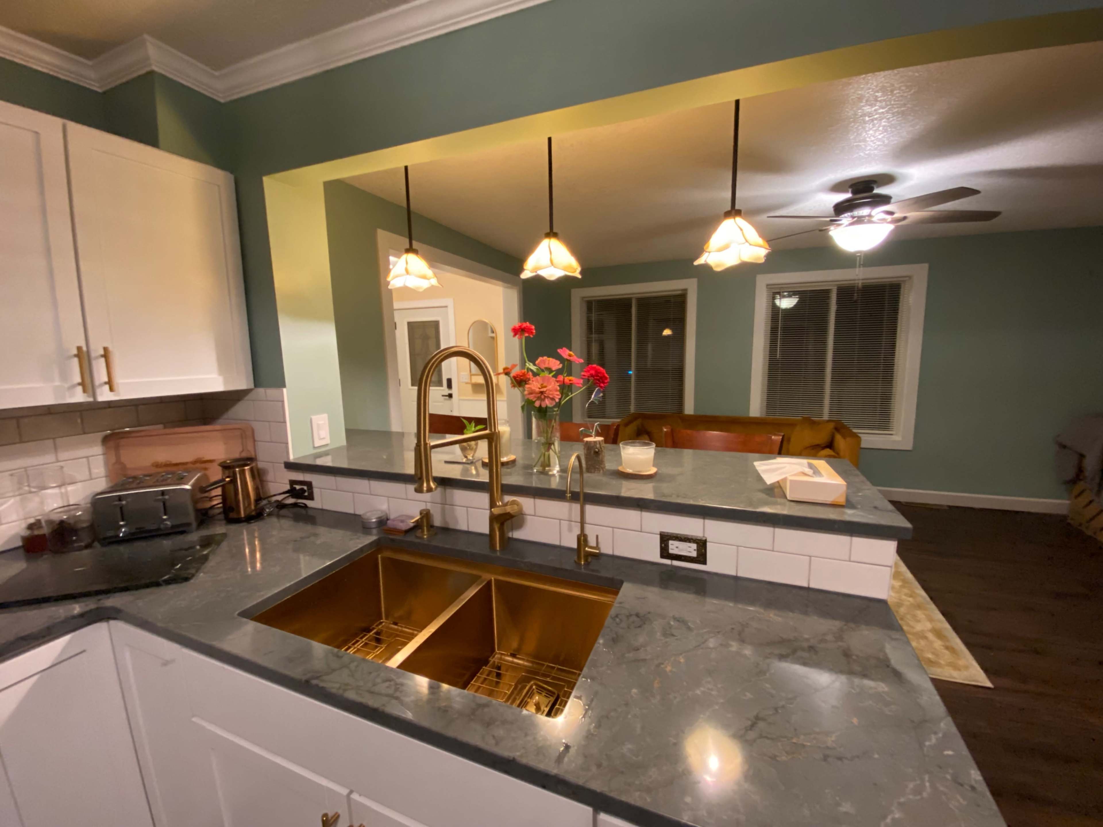 A modern kitchen features a double sink with a gold faucet and flowers on the countertop, while a living area with a brown sofa is visible in the background.