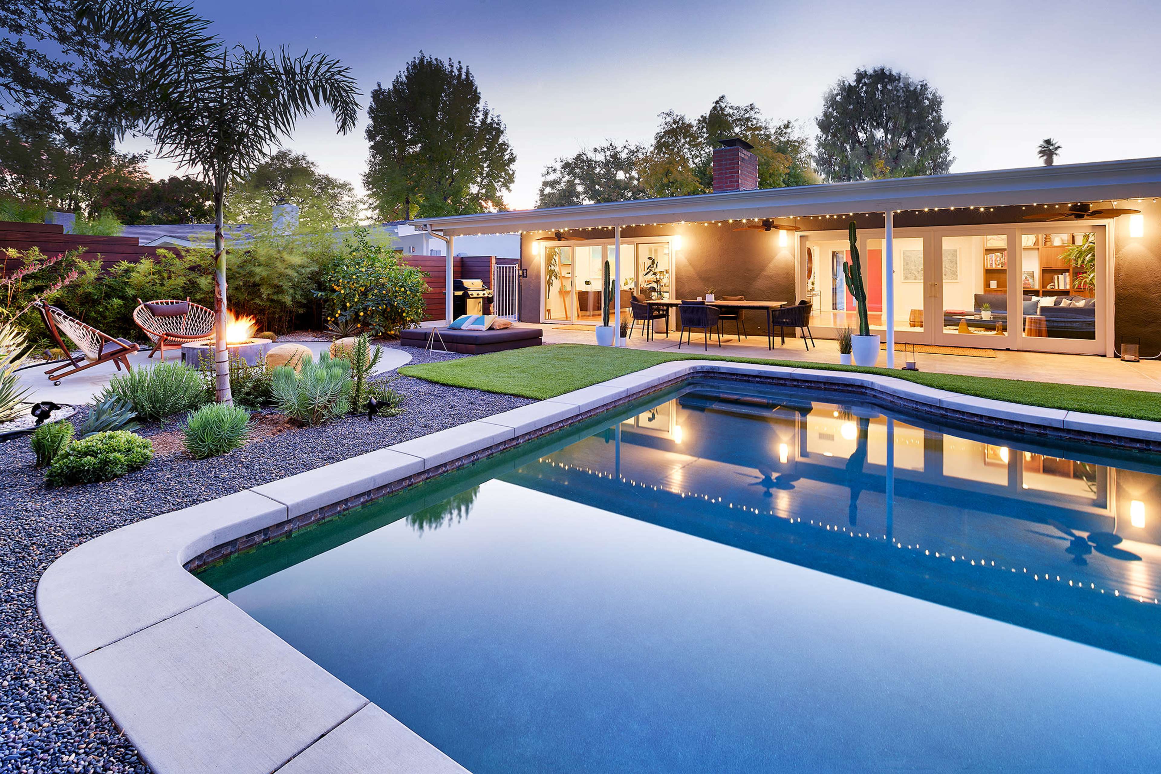 The image shows a modern backyard with a swimming pool, surrounded by landscaped greenery and a patio area featuring outdoor furniture and lighting.