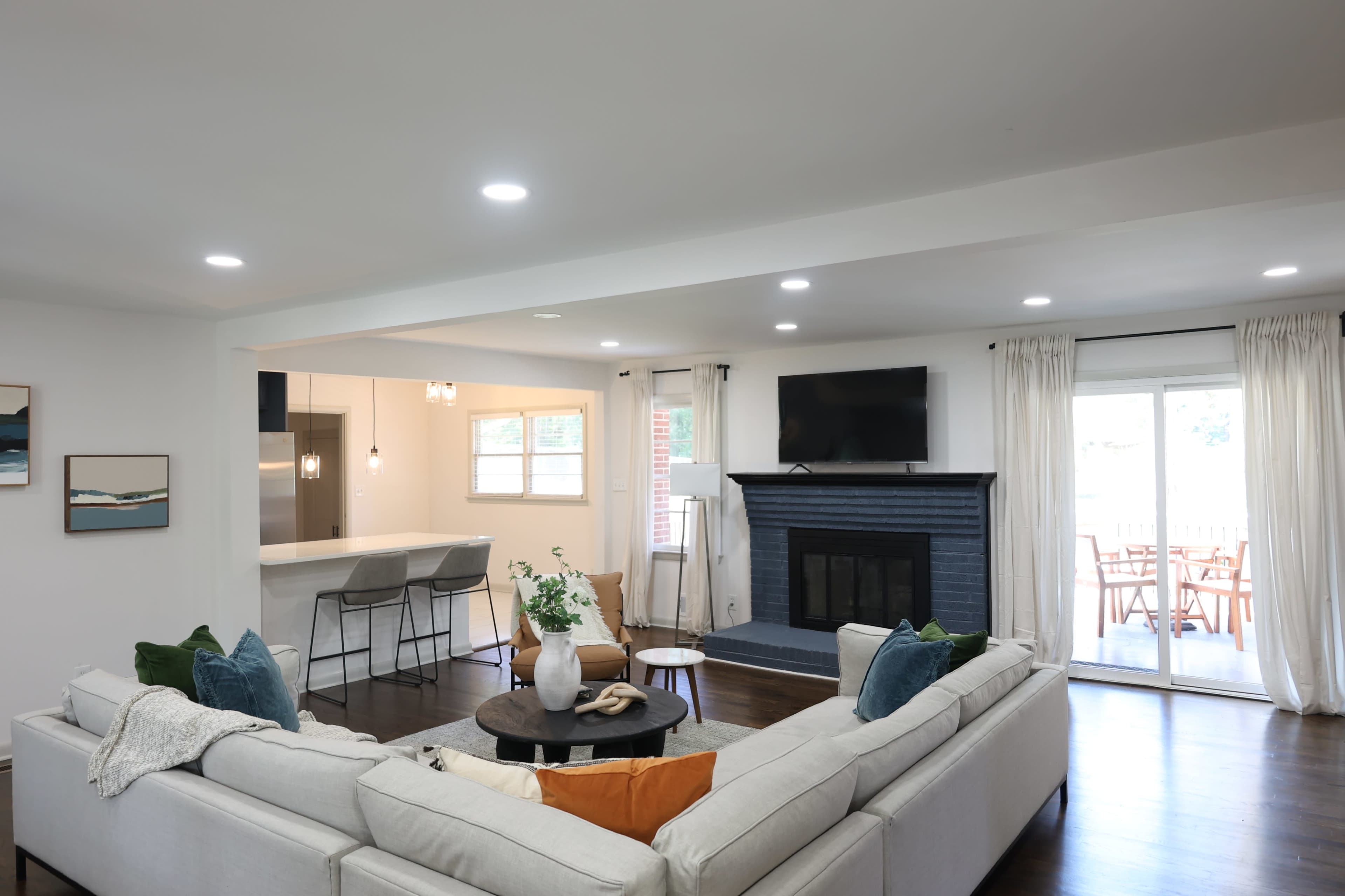 A modern living room with a sectional sofa, a coffee table, a fireplace, and a flat-screen TV, leading to an outdoor space with a dining area.