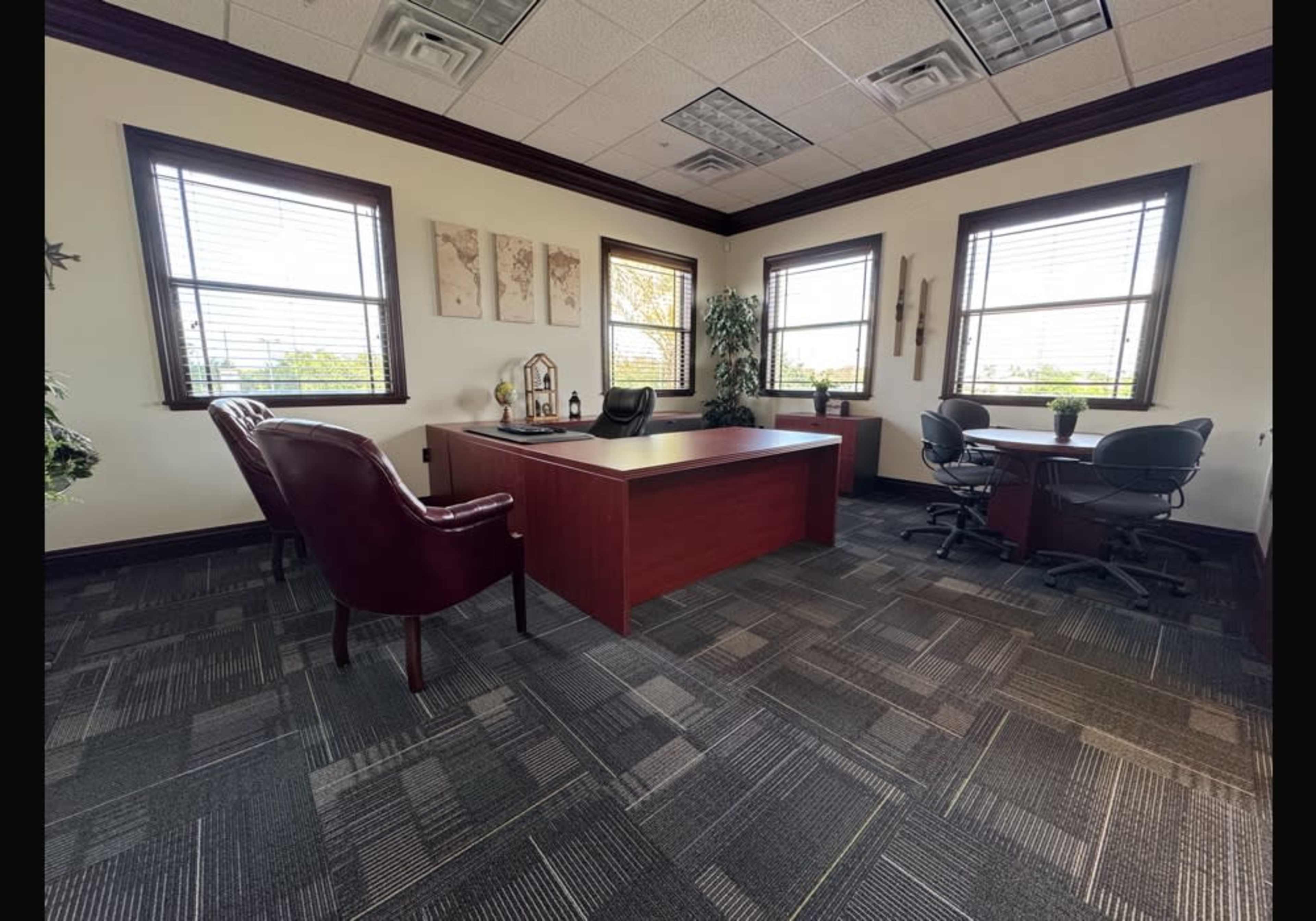 The image shows a well-furnished office space featuring an executive desk, two chairs, and a small meeting table with chairs, surrounded by large windows.