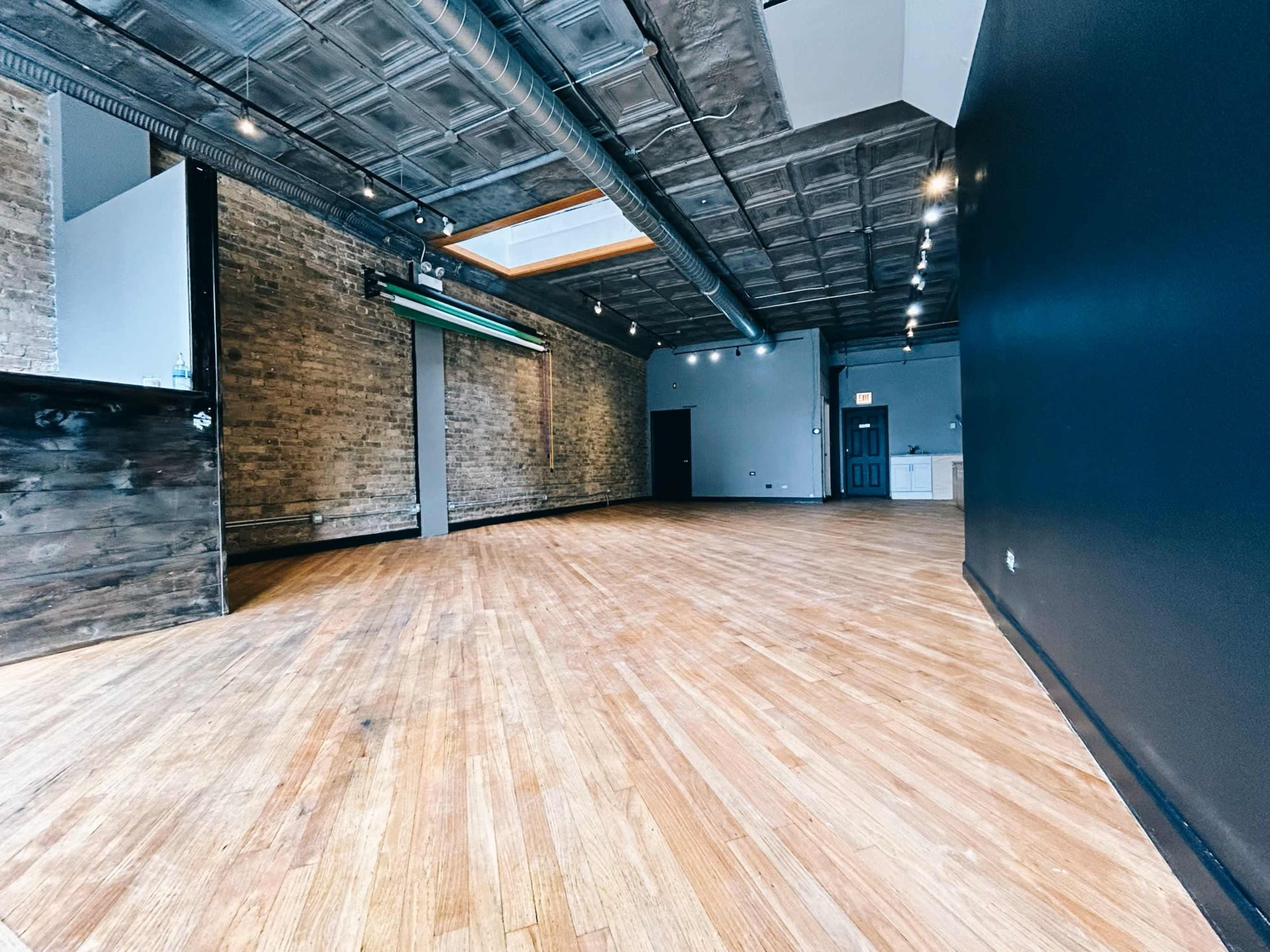 The image shows an empty room with hardwood flooring, exposed brick walls, and a black accent wall, featuring overhead lighting and a bar area on one side.