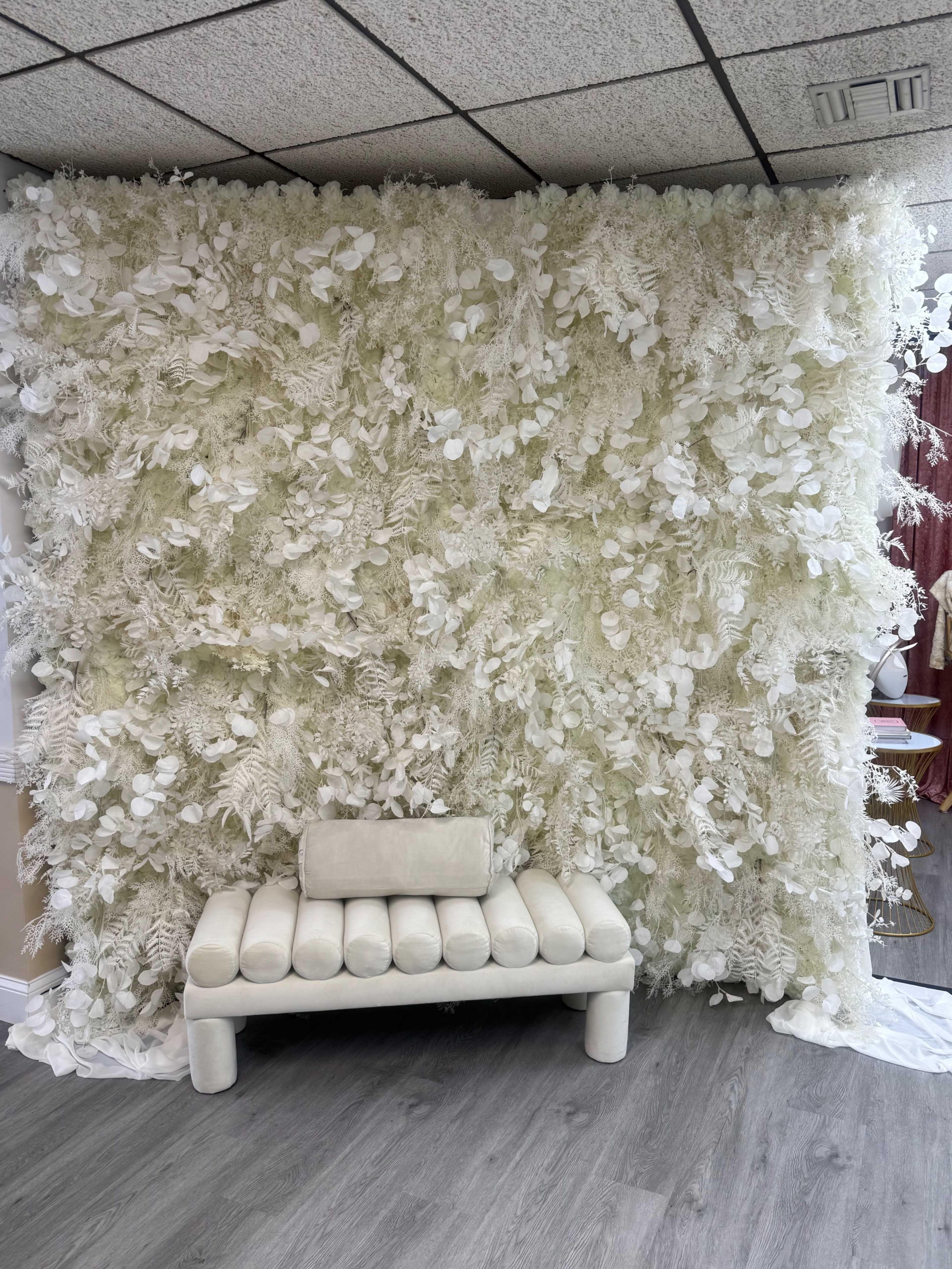 The image shows a textured white wall made of artificial foliage, behind a simple, light-colored bench in a room with a wooden floor.