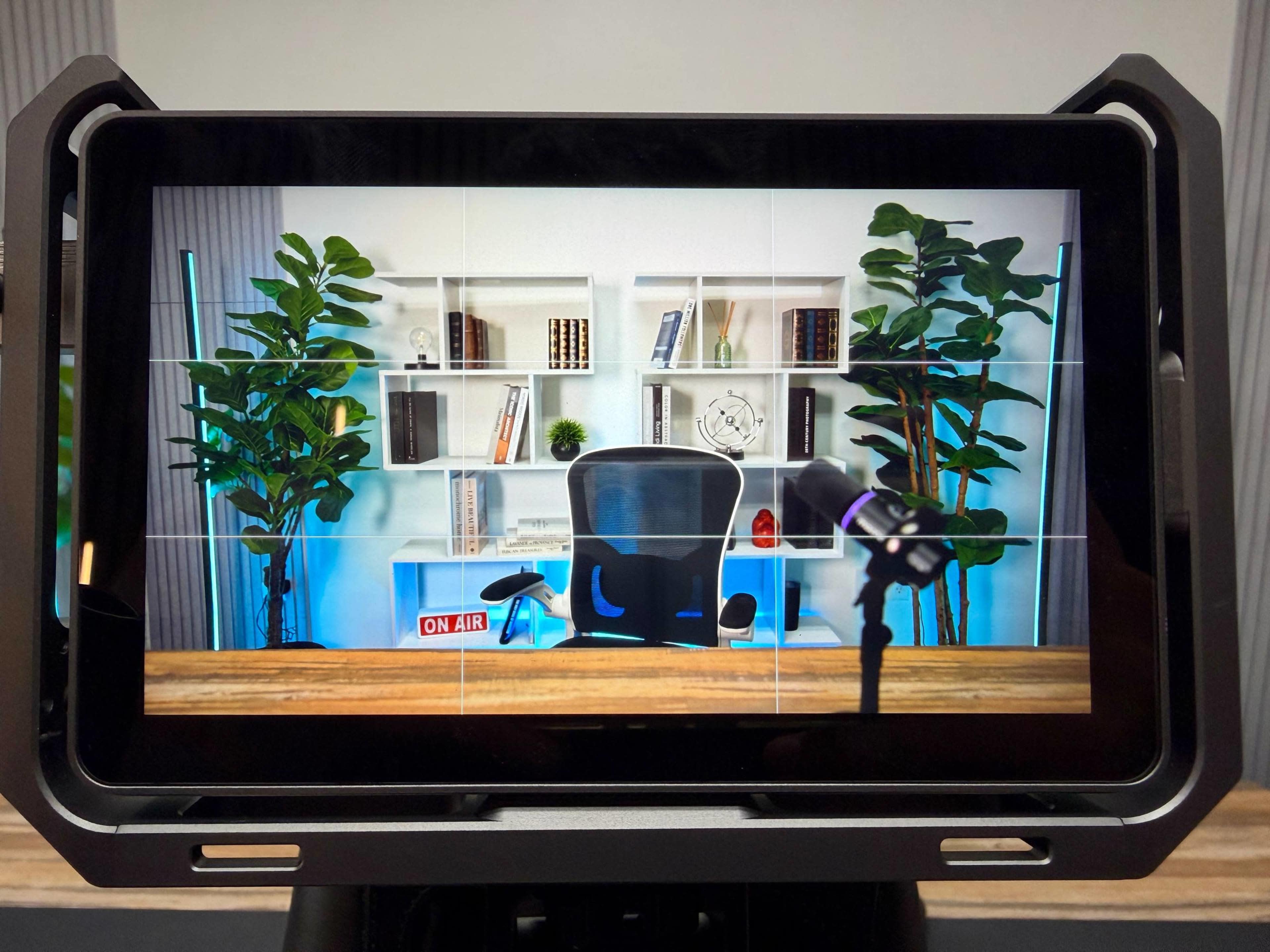 A tablet display shows a well-organized studio setup with a black office chair, a microphone, and shelves filled with books and decorative items, flanked by green plants.