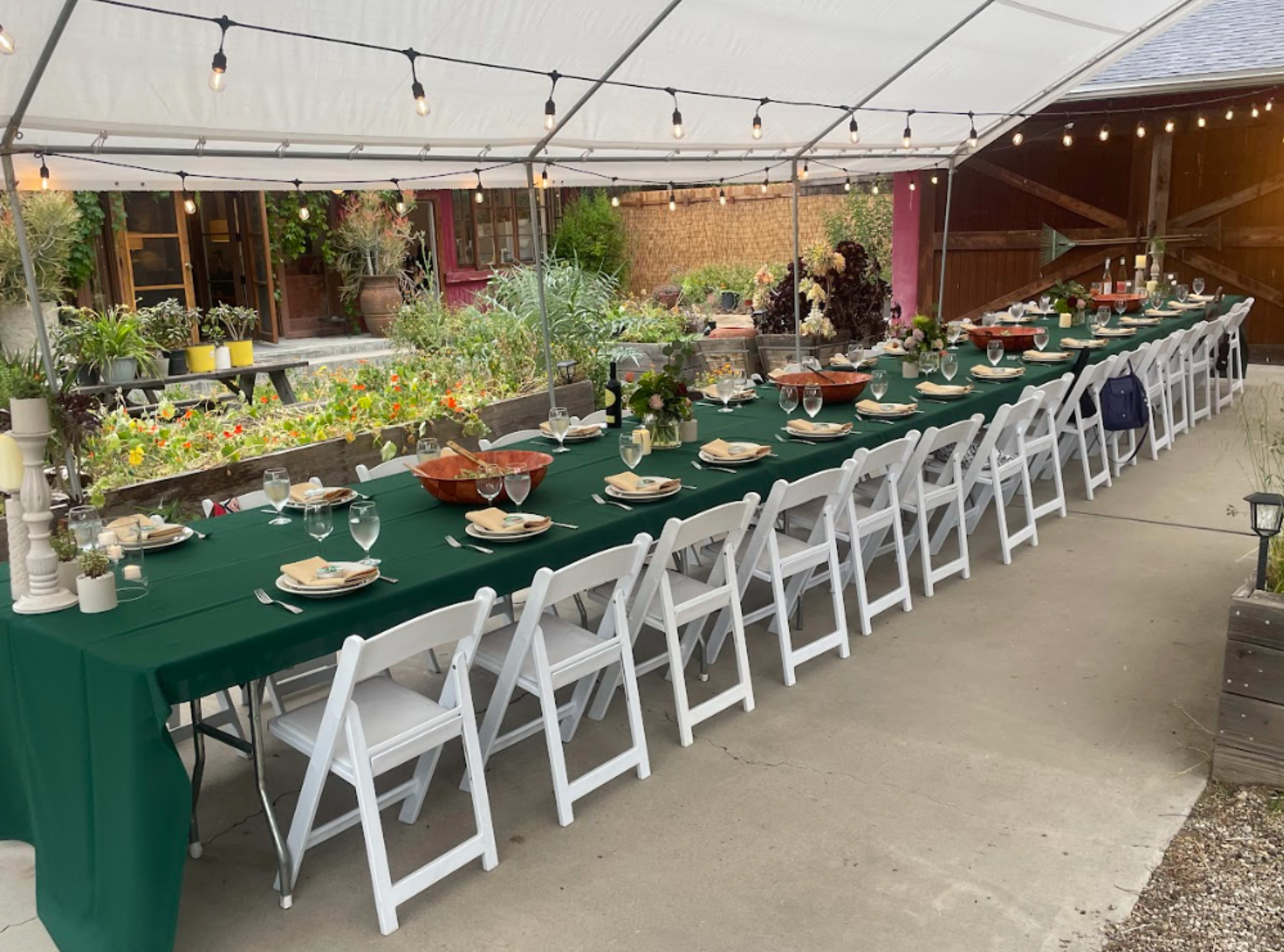 A long rectangular table is set with green tablecloths and white chairs under a tent, surrounded by plants and decorative lighting.