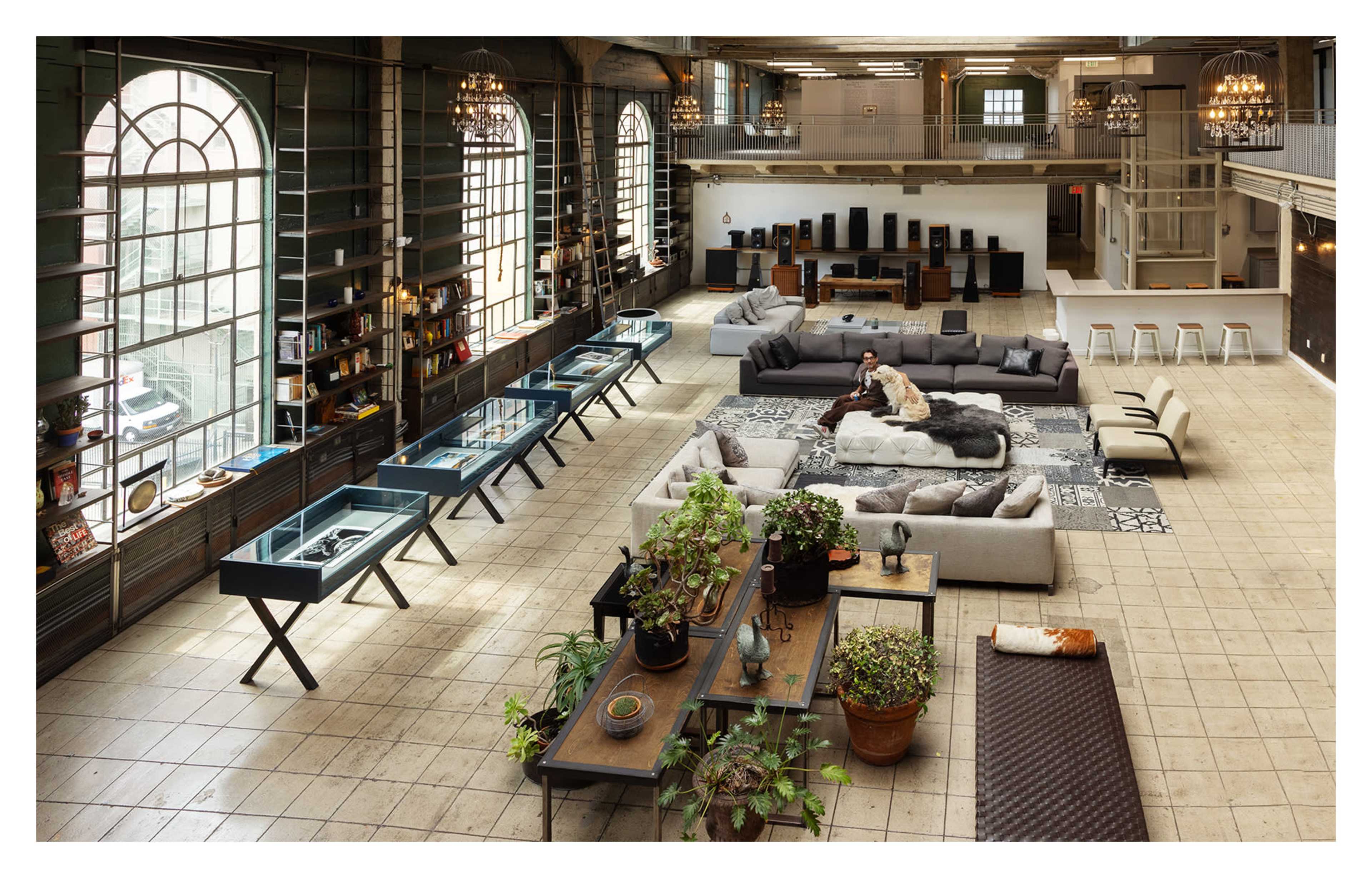 The image shows a spacious, modern loft with high ceilings, large windows, and distinct areas for seating and displays surrounded by greenery.