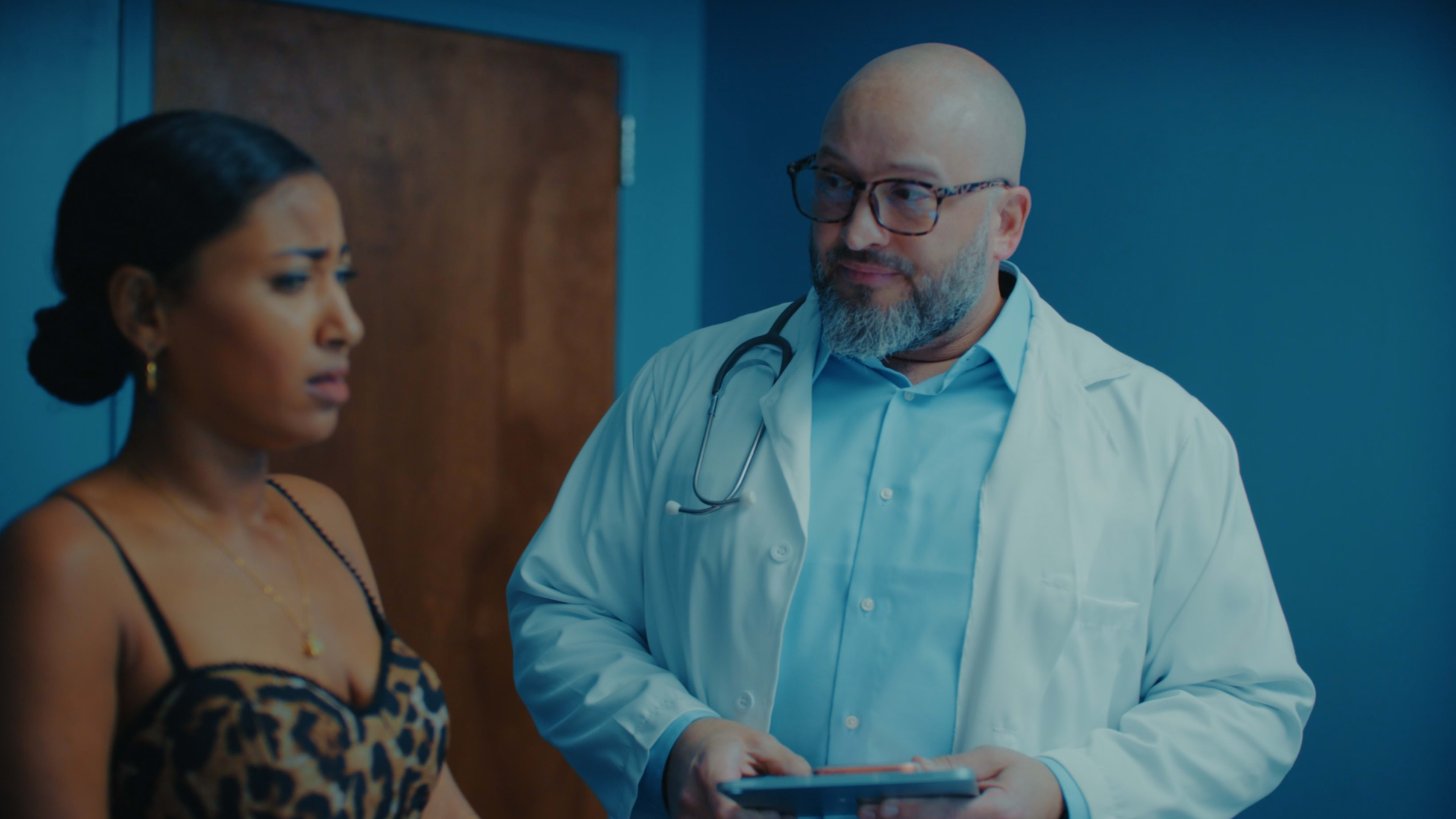 A woman in a patterned dress stands next to a man in a white coat and glasses, who is holding a tablet in a medical office.