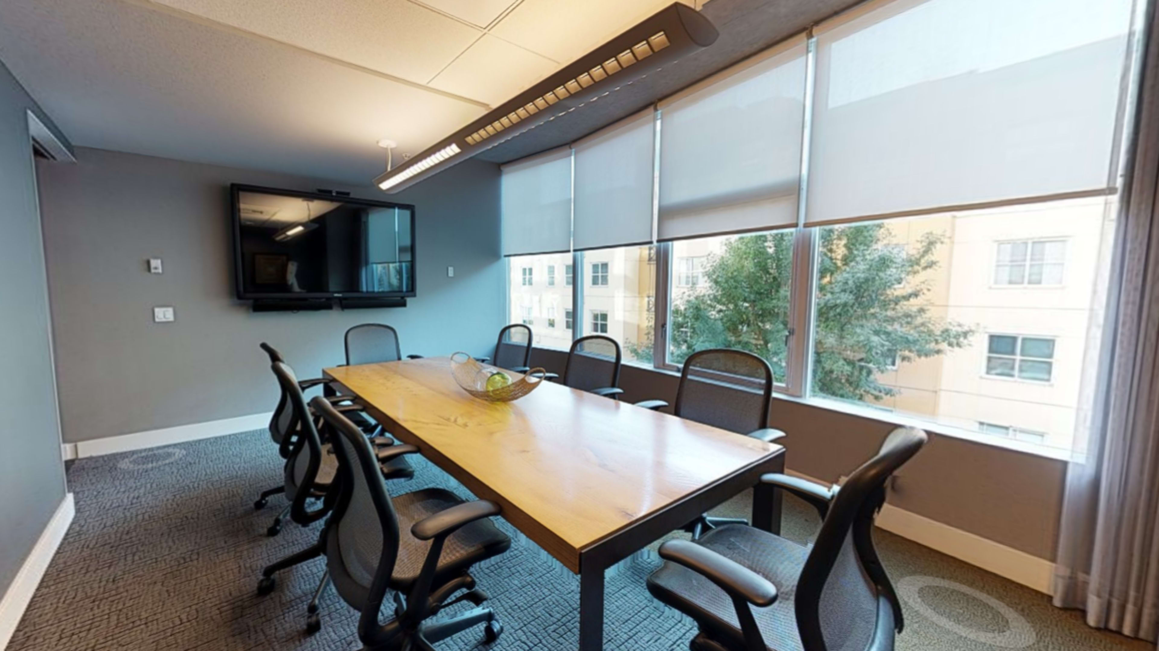 A conference room features a large wooden table surrounded by eight black mesh chairs, with a wall-mounted TV and large windows overlooking a building exterior.