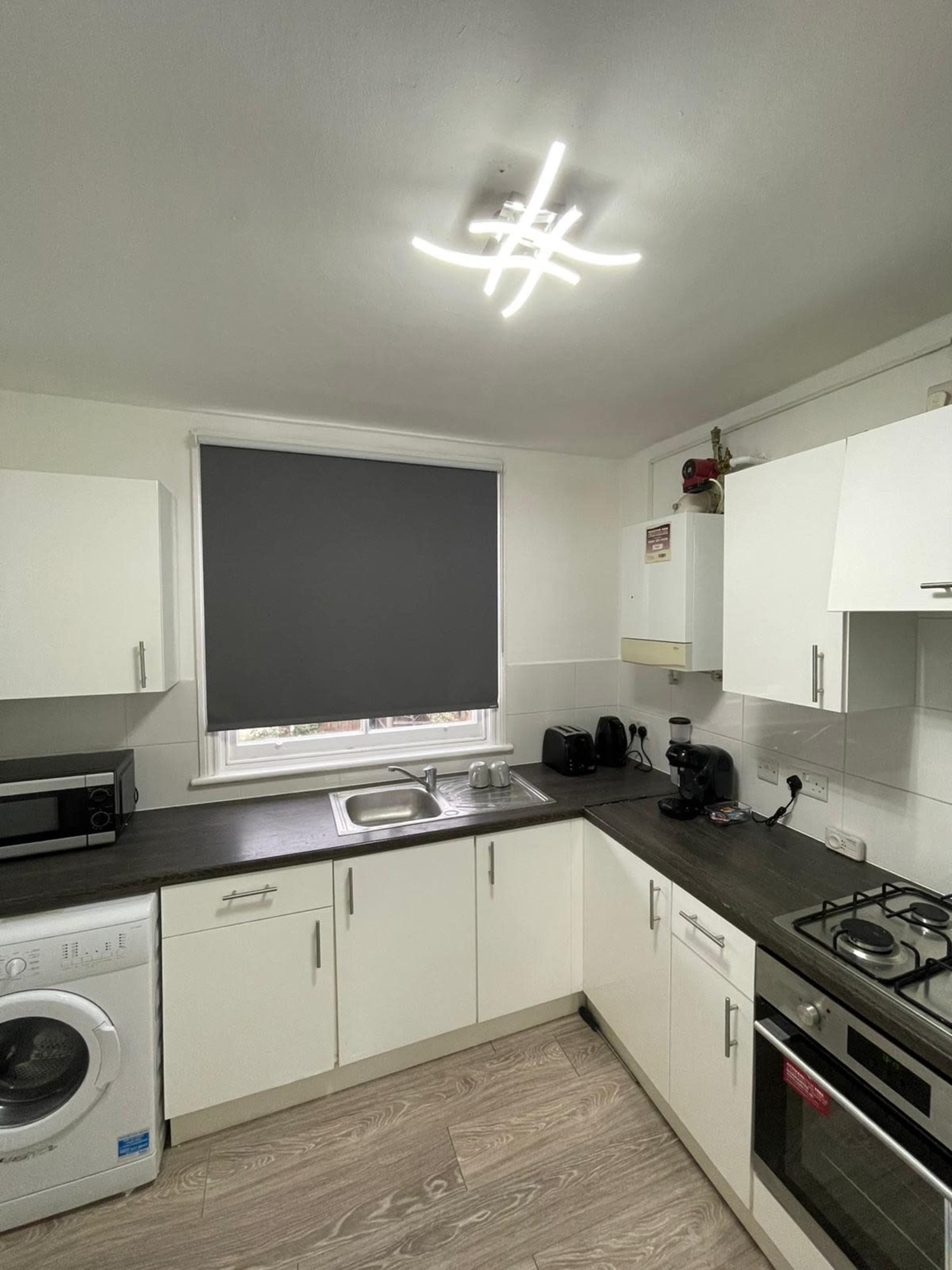 A modern kitchen featuring white cabinetry, a stainless steel sink, a washing machine, and a gray roller blind.