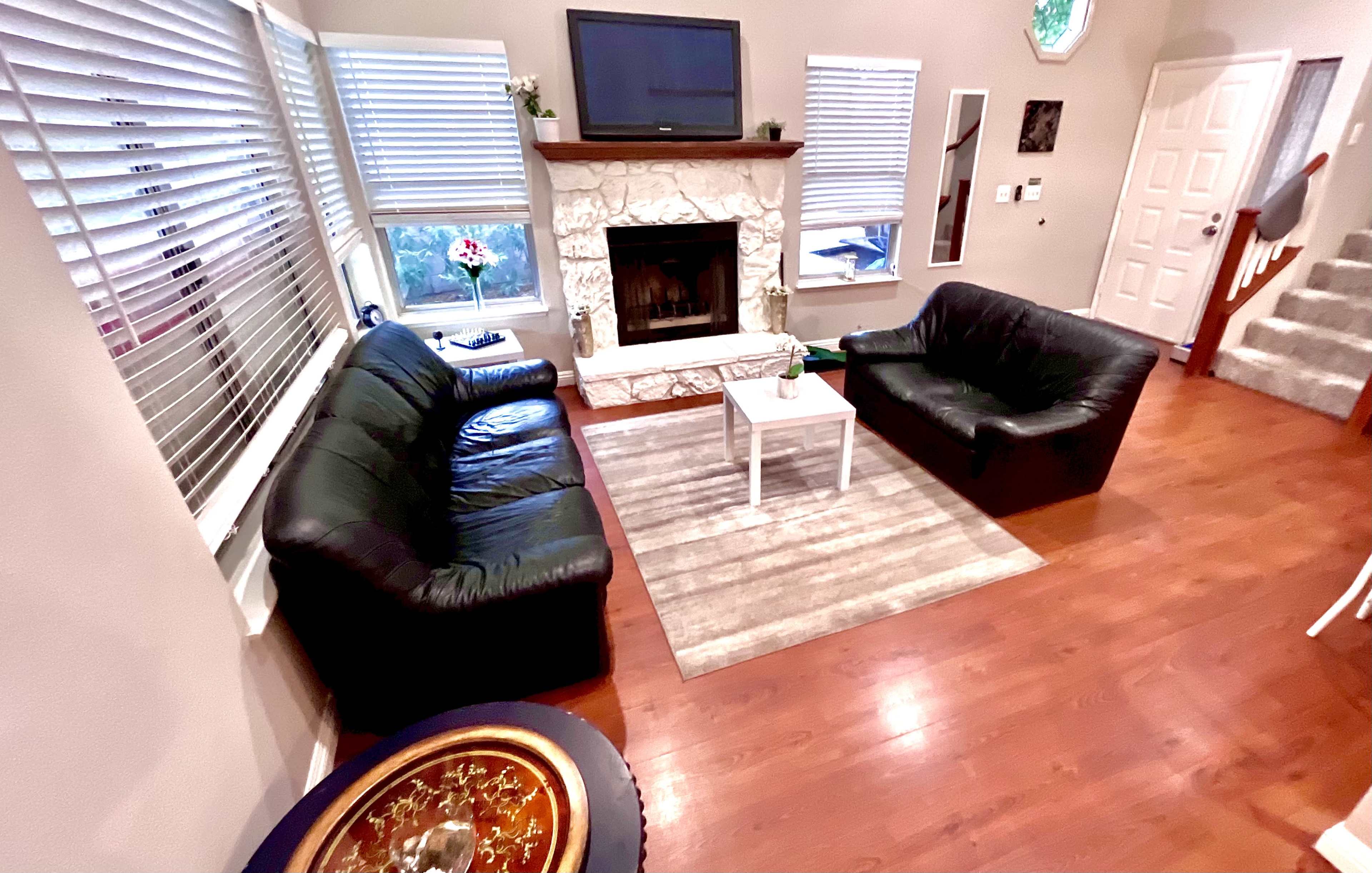 A living room features two black leather sofas facing a small white table, with a stone fireplace and a television mounted above it.