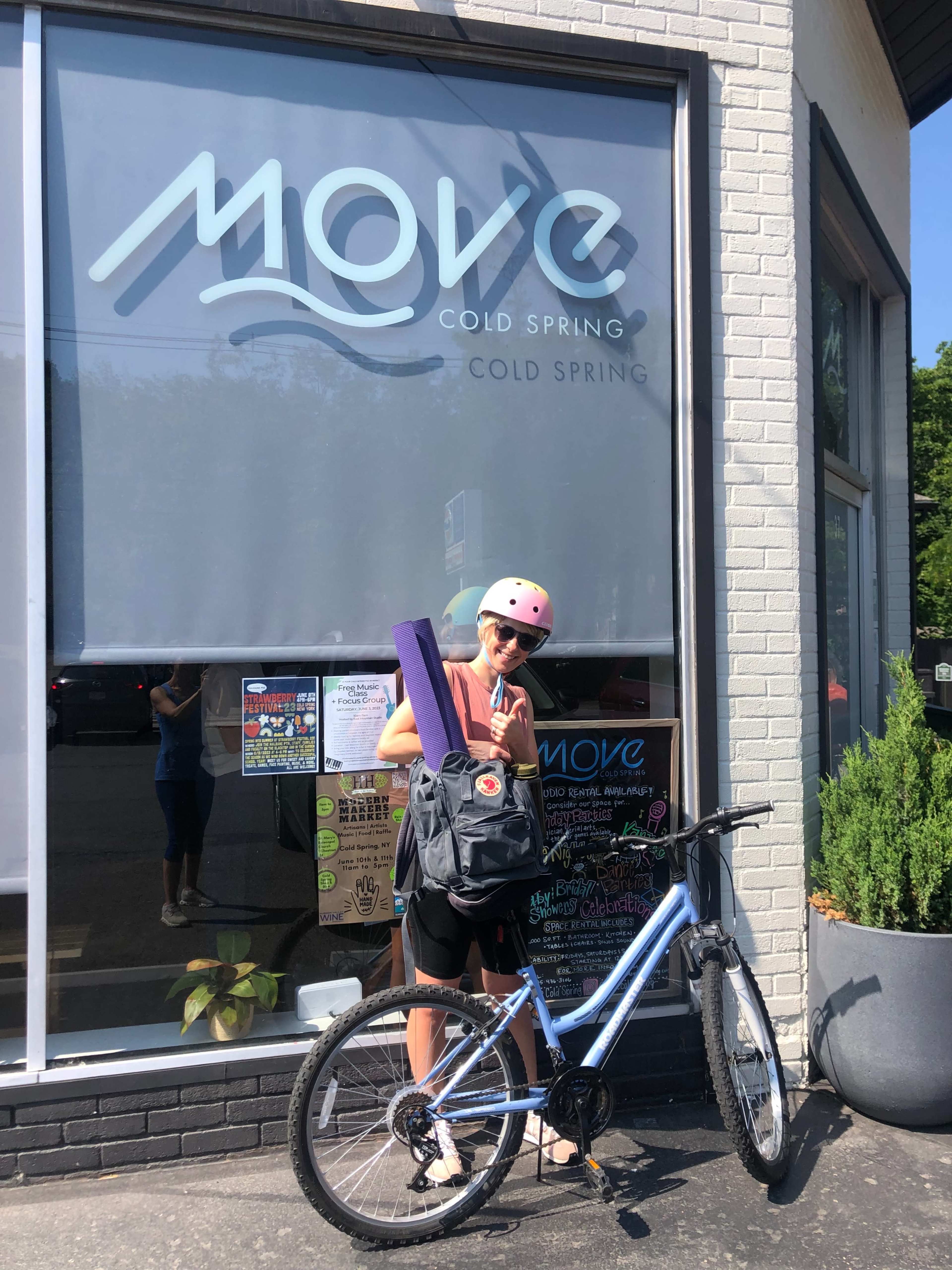 A person wearing a helmet stands next to a bicycle with a yoga mat and backpack in front of a business called "MOVE COLD SPRING."