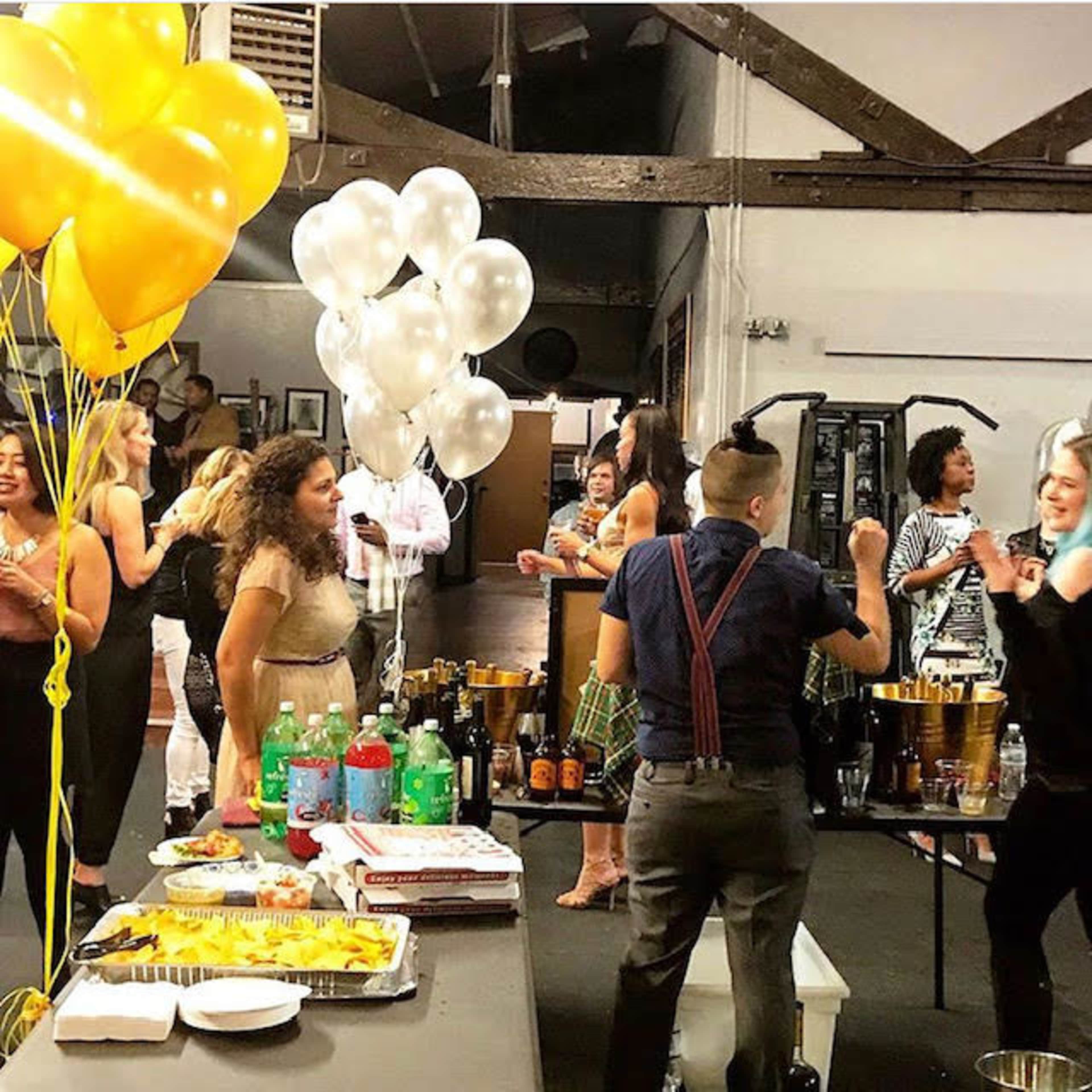 A festive gathering takes place in a venue with guests dancing, colorful balloons, and a table of food and drinks.