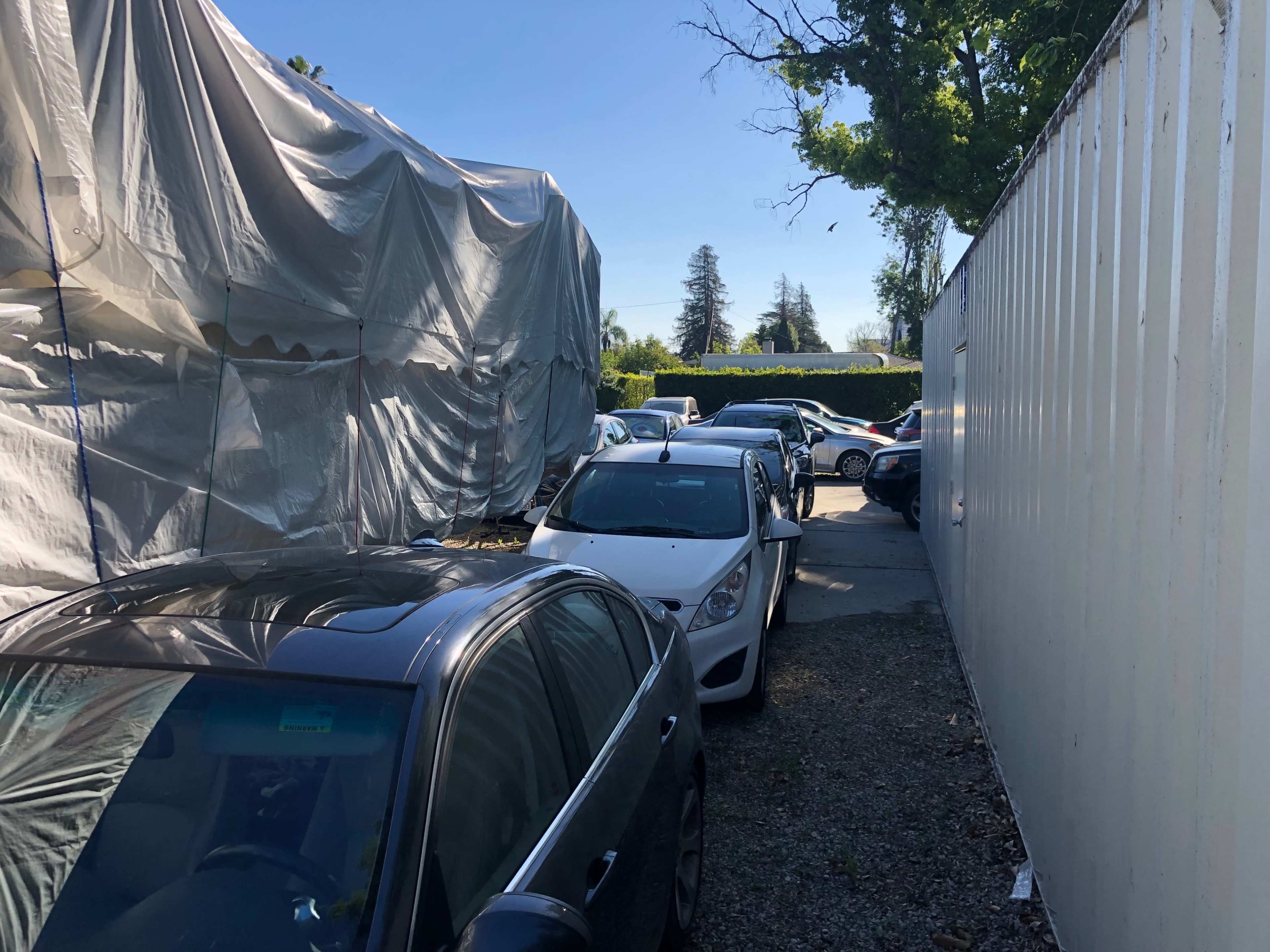 The image shows a narrow pathway between parked cars and a covered structure, with additional vehicles in the background under clear blue skies.
