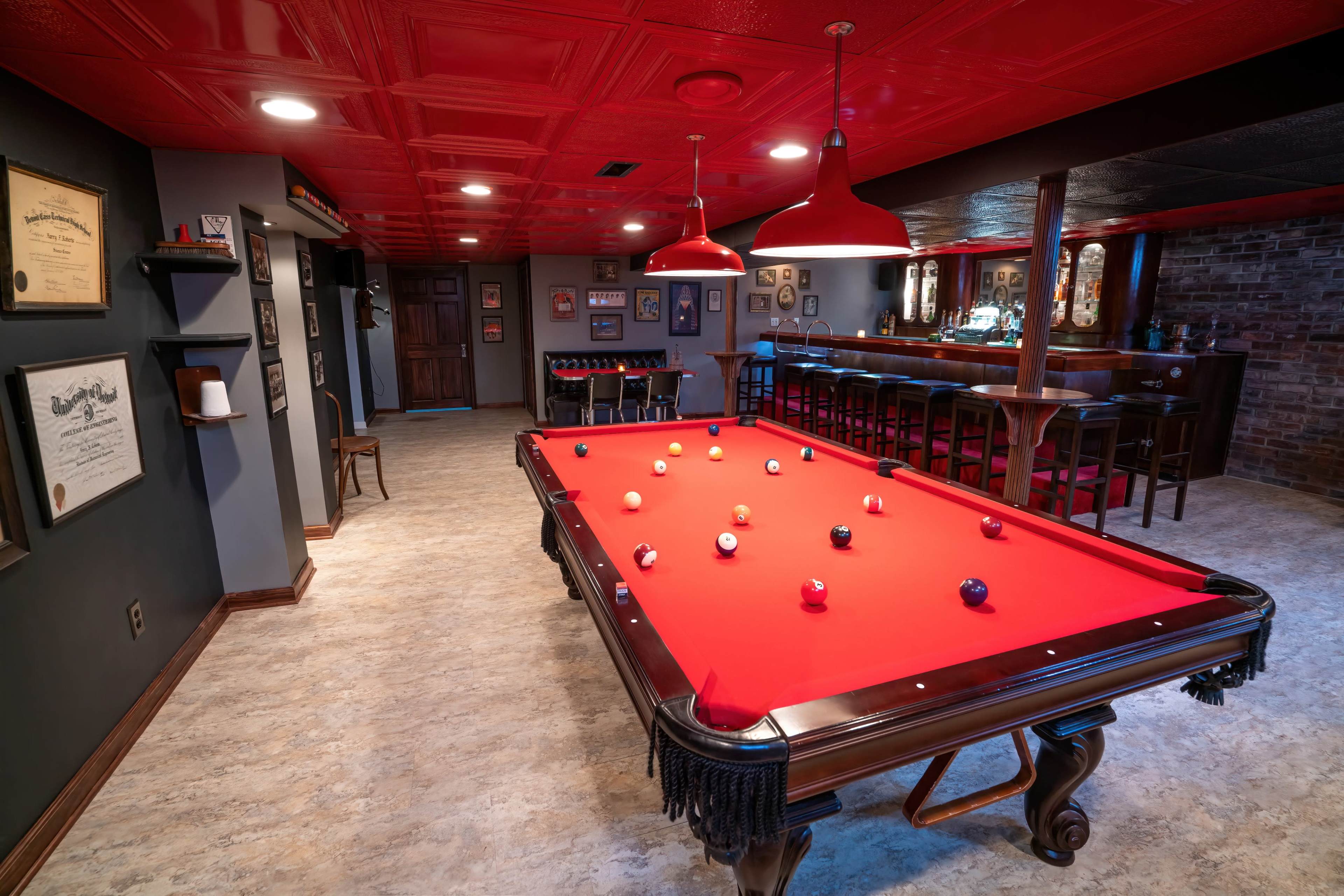 A basement game room featuring a red-felt pool table surrounded by barstools and wall decorations.