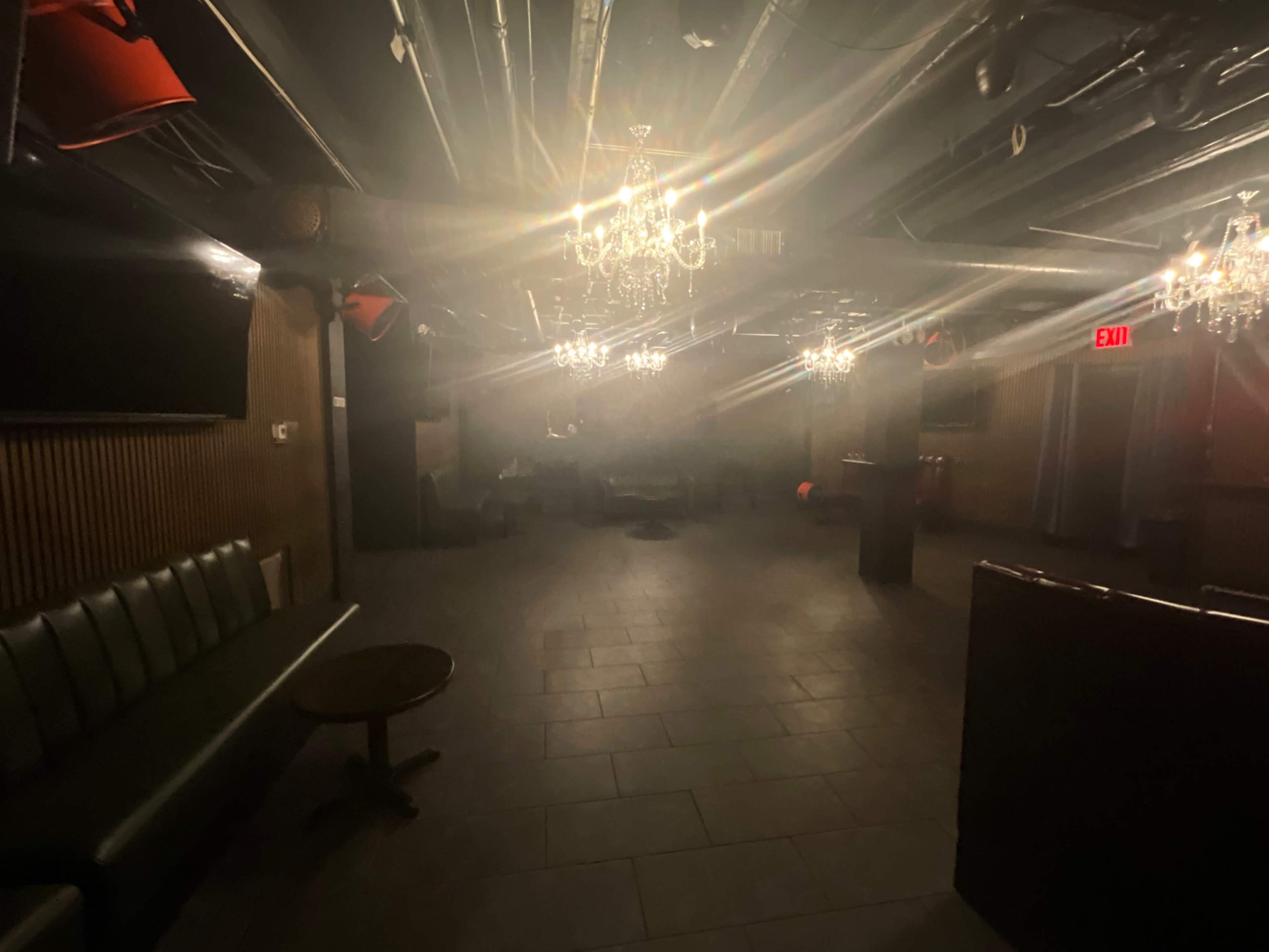 The image shows a dimly lit room with chandeliers hanging from the ceiling and a few scattered pieces of furniture.