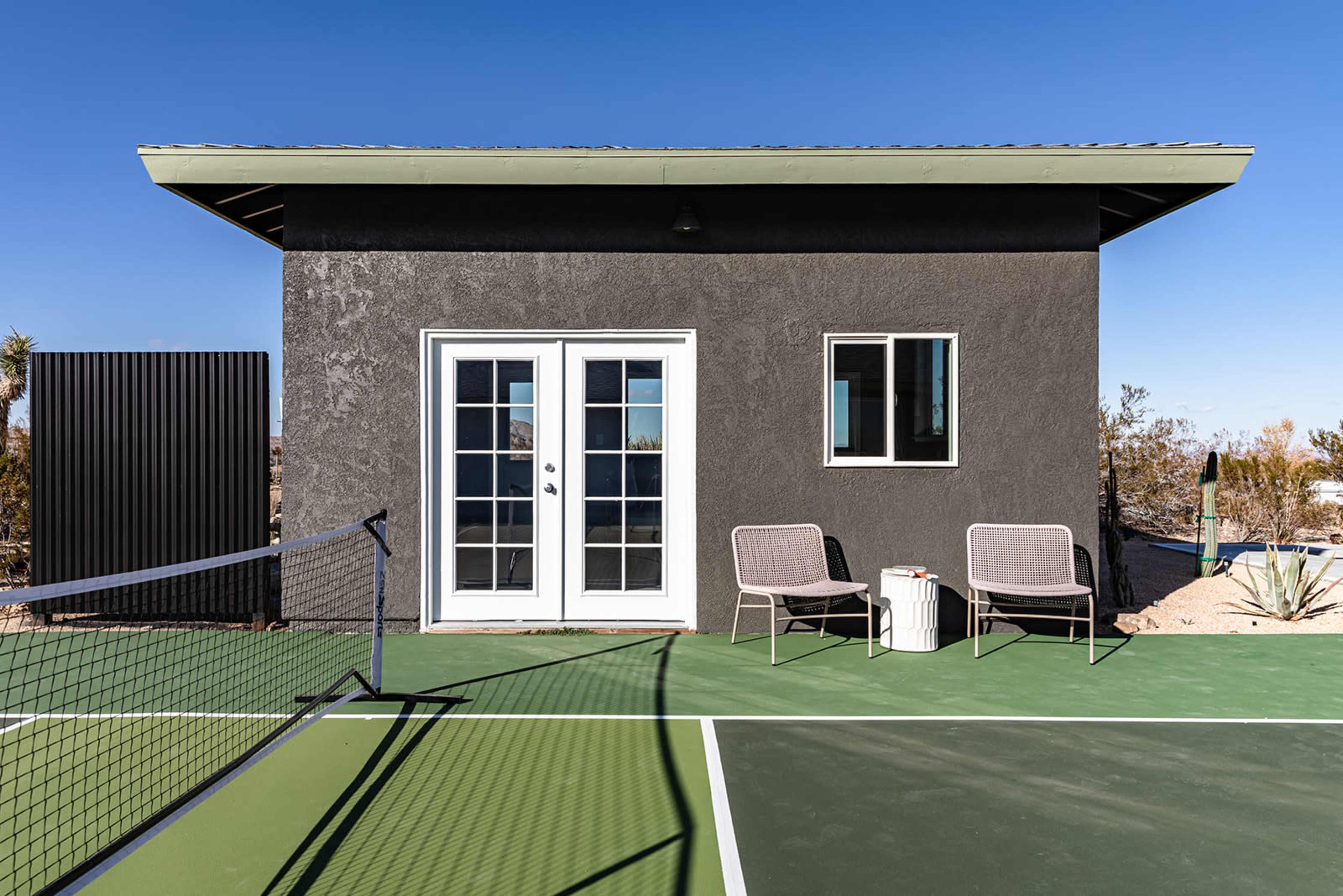 The image shows a modern small building with a gray exterior, featuring French doors and two chairs, situated next to a green tennis court.