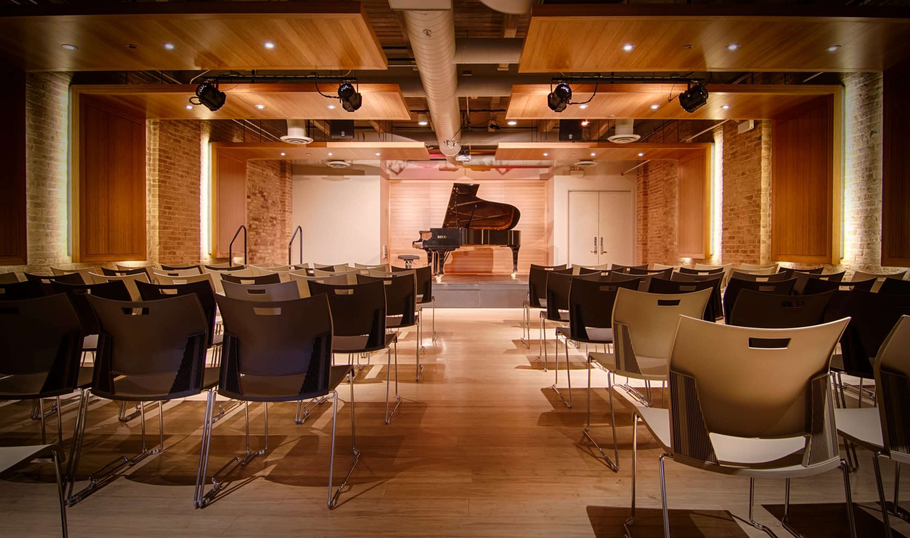 A concert hall with rows of chairs facing a grand piano on stage, surrounded by wooden beams and warm lighting.