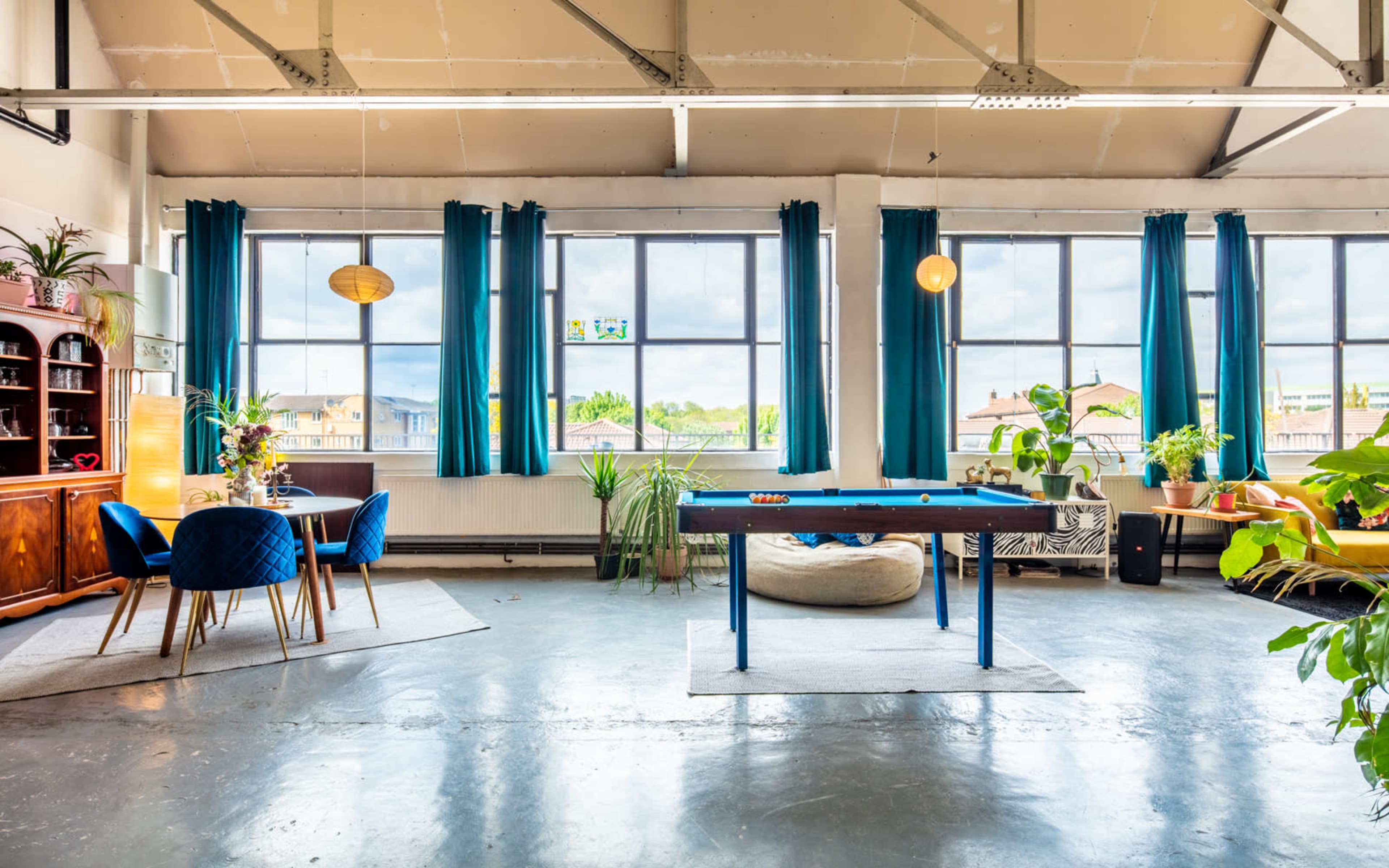 A spacious room features large windows, a pool table, plants, and various seating arrangements.