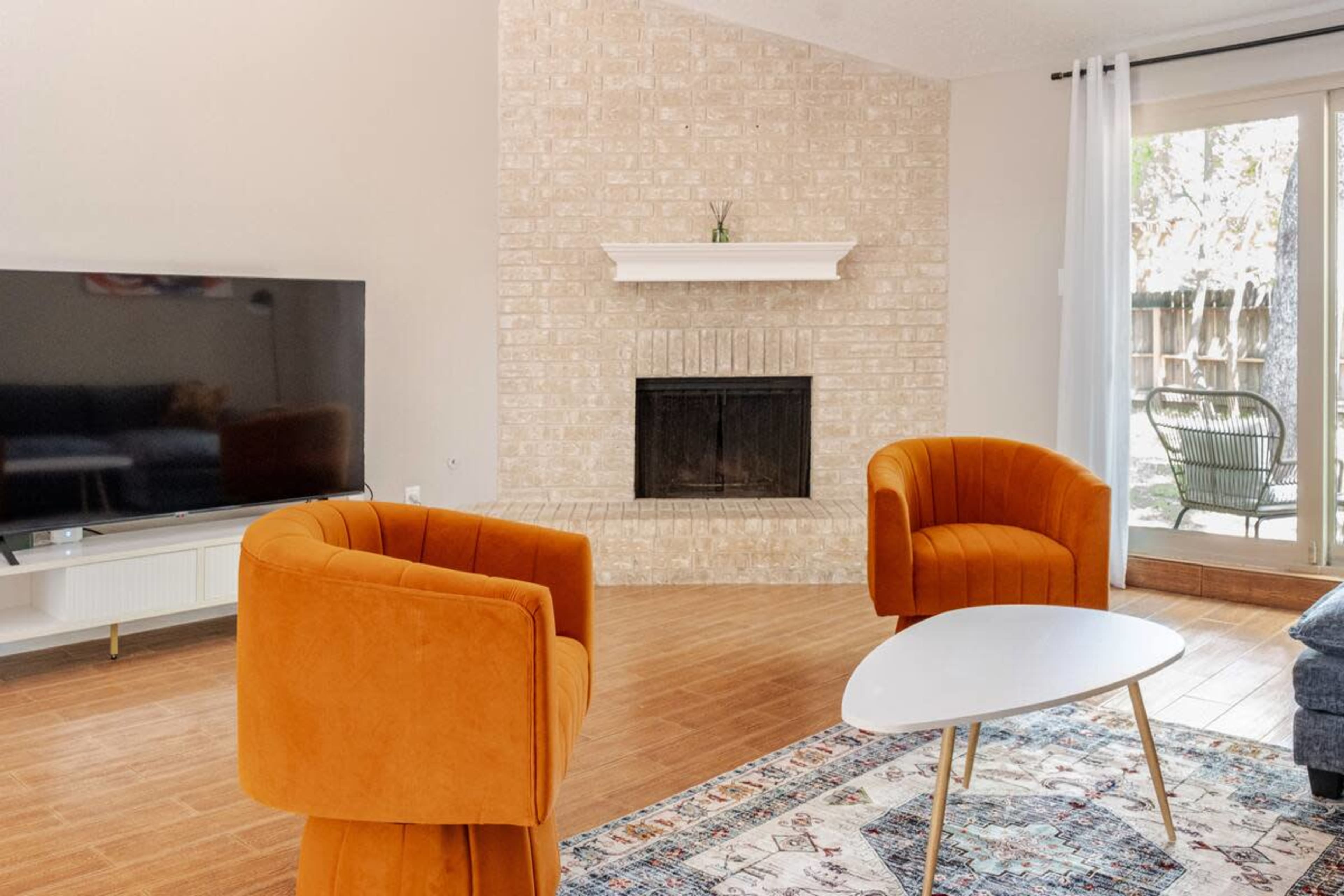 The living room features two orange velvet chairs facing a white coffee table, with a television and a brick fireplace on one wall, and large sliding doors leading to an outdoor space.