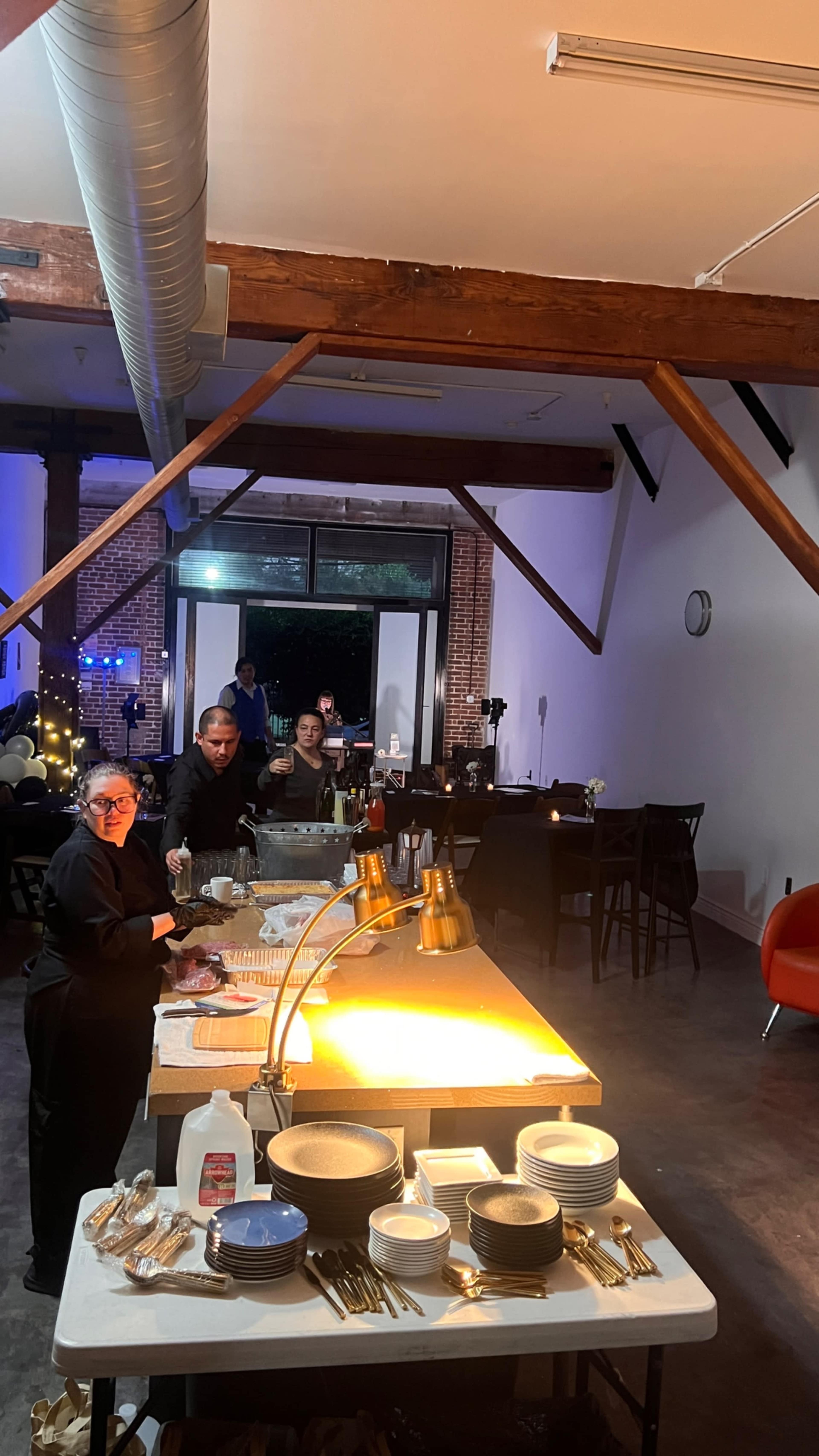 A spacious dining area is set up with a buffet table at the front, where two staff members are preparing food, while guests can be seen seated in the background.