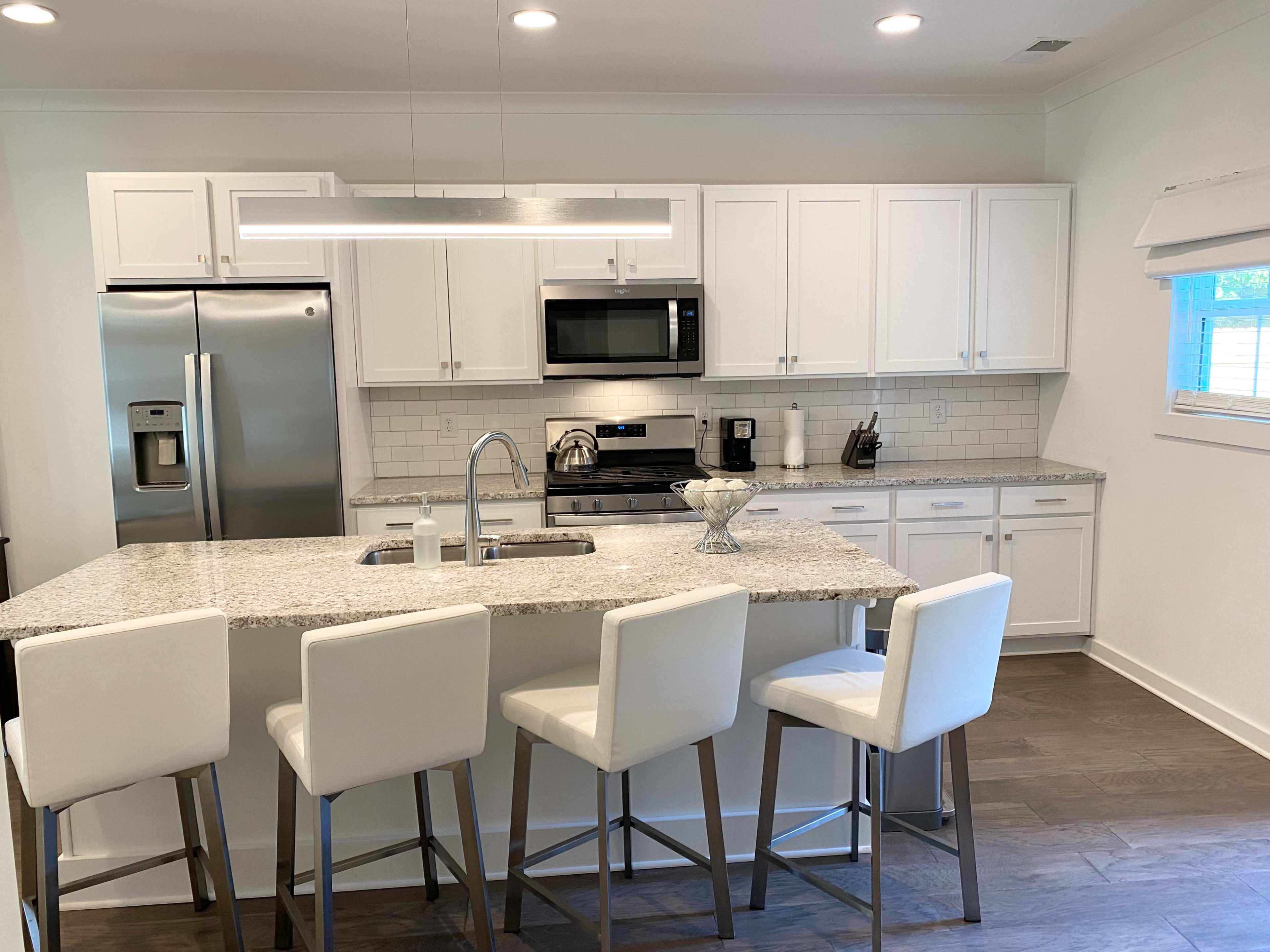 A modern kitchen features white cabinetry, stainless steel appliances, a large granite island with seating, and bright lighting.
