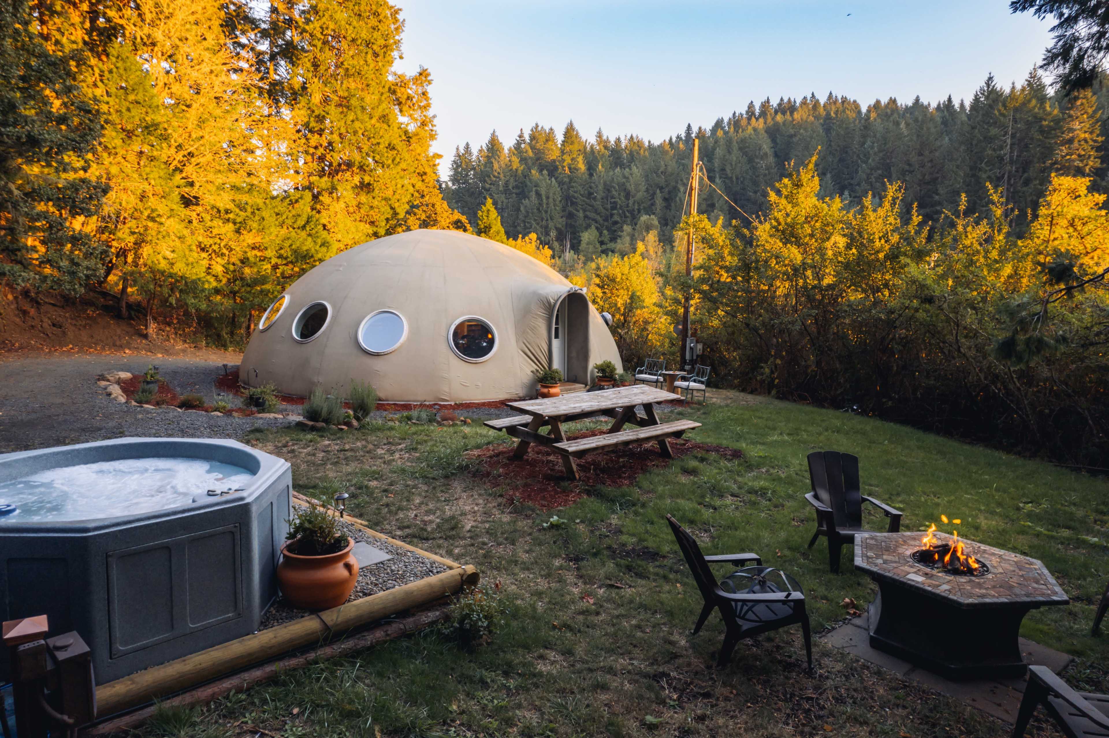 The image shows a geodesic dome cabin surrounded by autumn trees, with a hot tub, picnic table, and fire pit in the foreground.