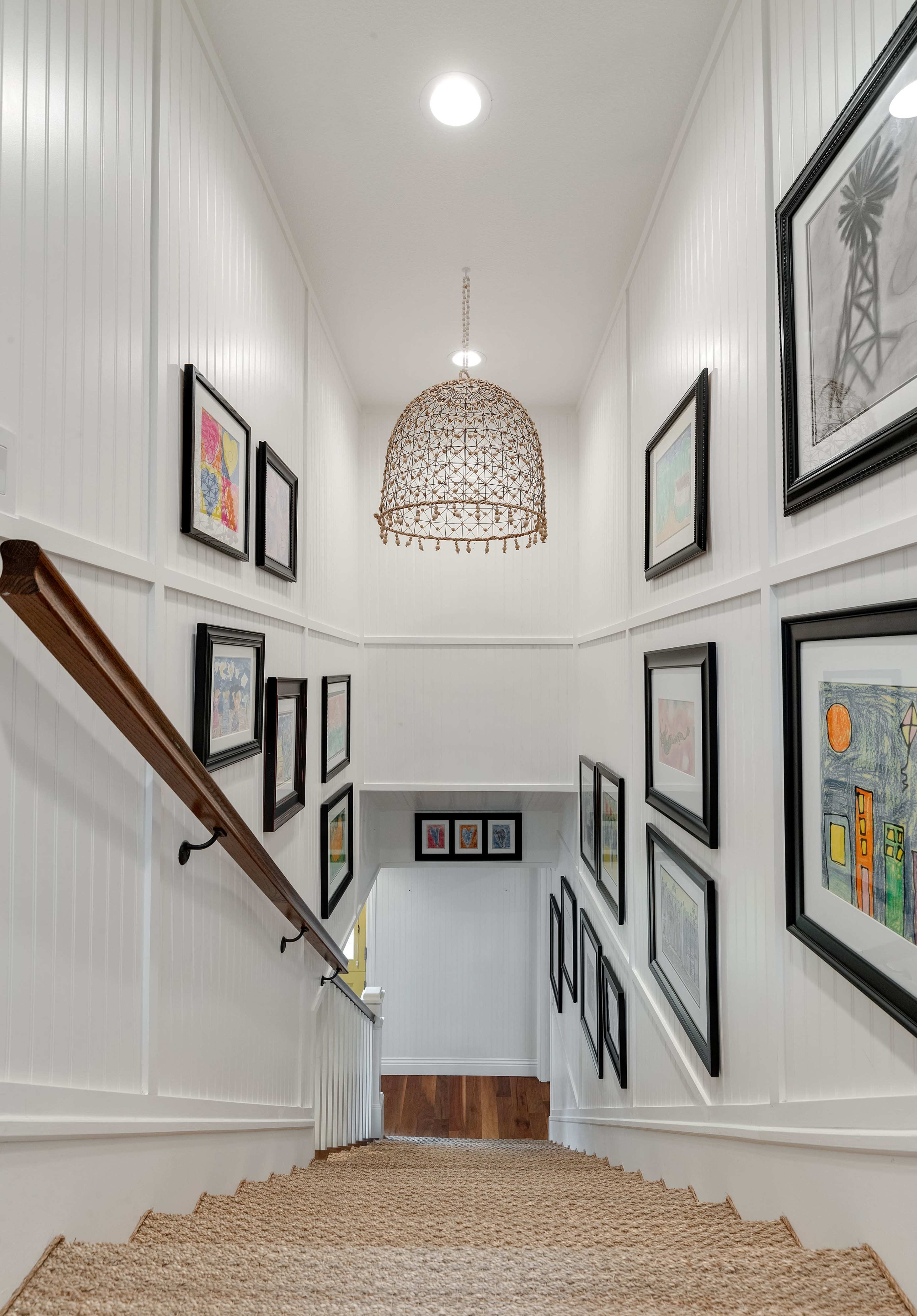 A well-lit staircase with a decorative chandelier, featuring framed artwork on the walls and a rug covering the steps.