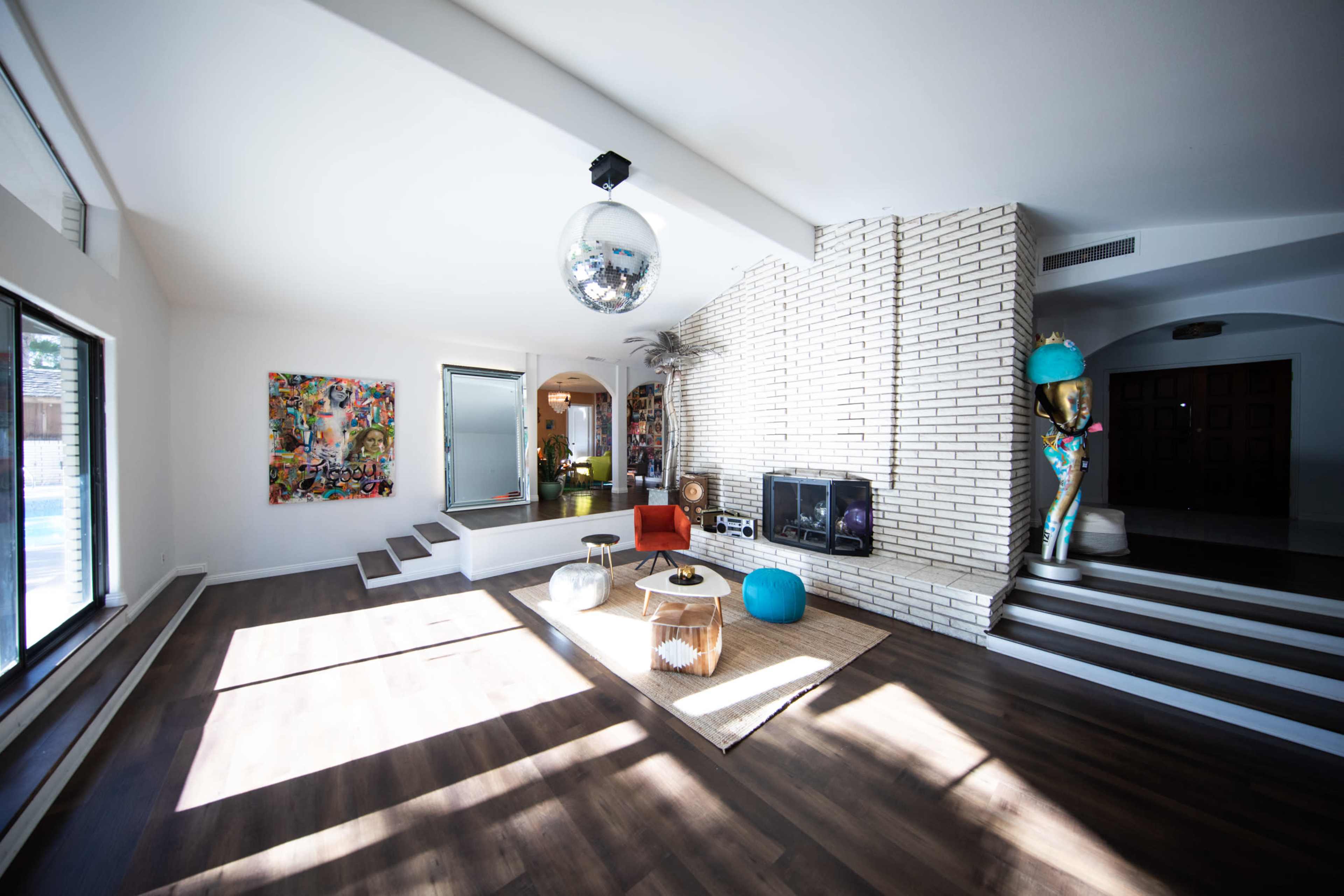 The image shows a spacious living room featuring large windows, a disco ball, colorful art on the walls, and a mix of modern furniture arranged around a central coffee table.