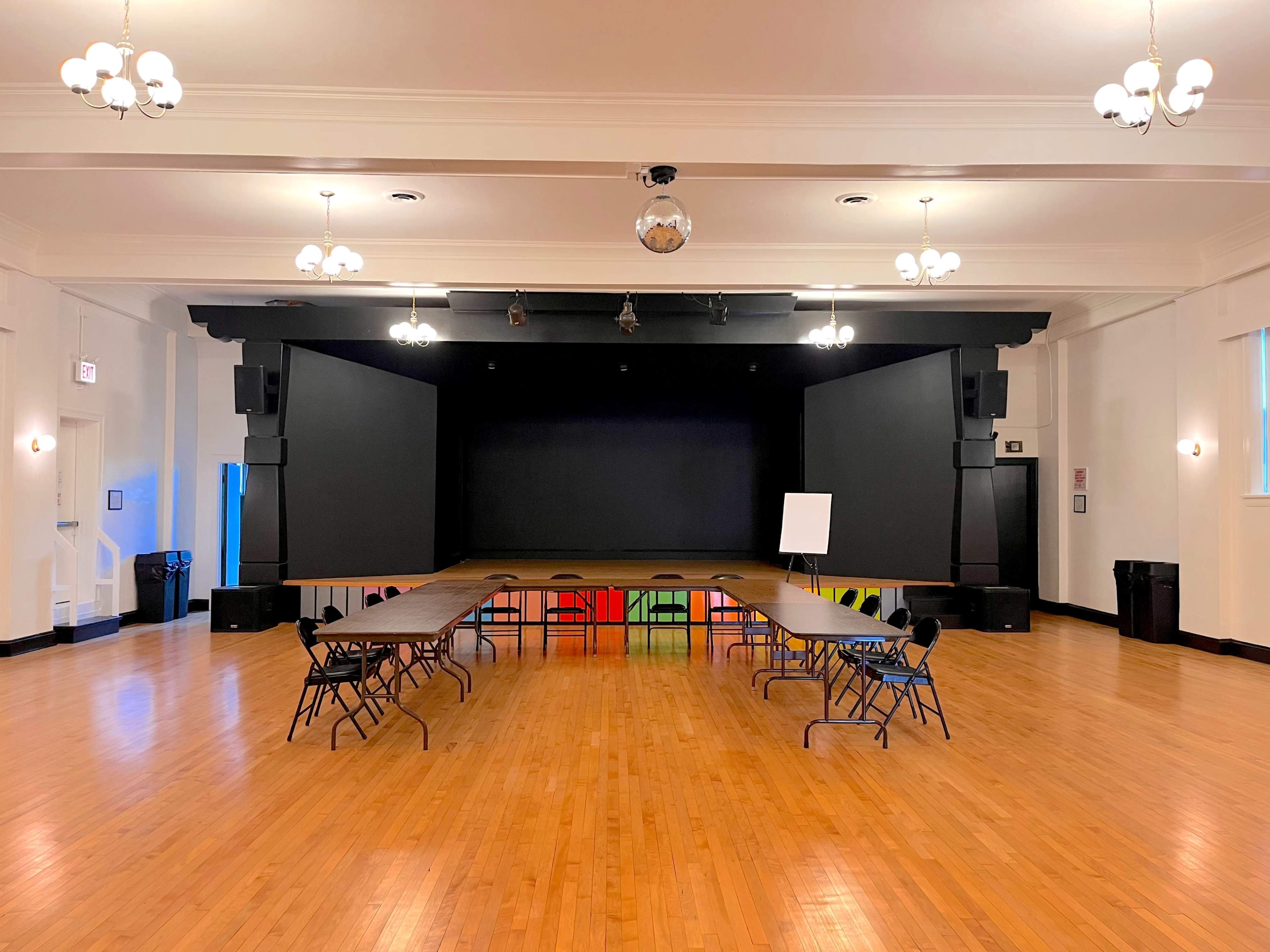 A spacious event hall with a stage at the front, featuring a row of tables arranged in a U-shape and chairs facing the stage.