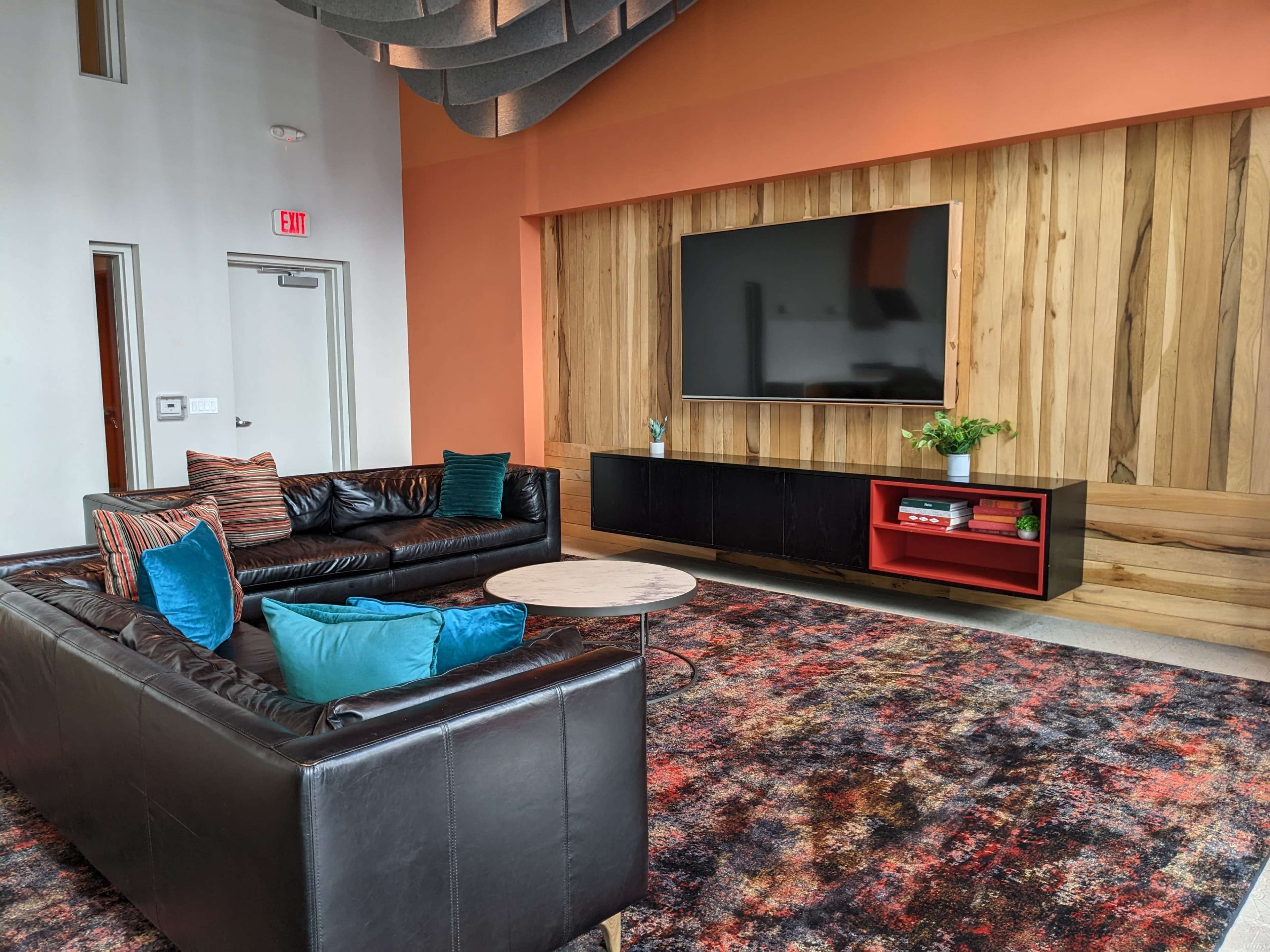 The room features a leather sofa set, a round coffee table, a large television mounted on a wooden accent wall, and colorful decorative elements.