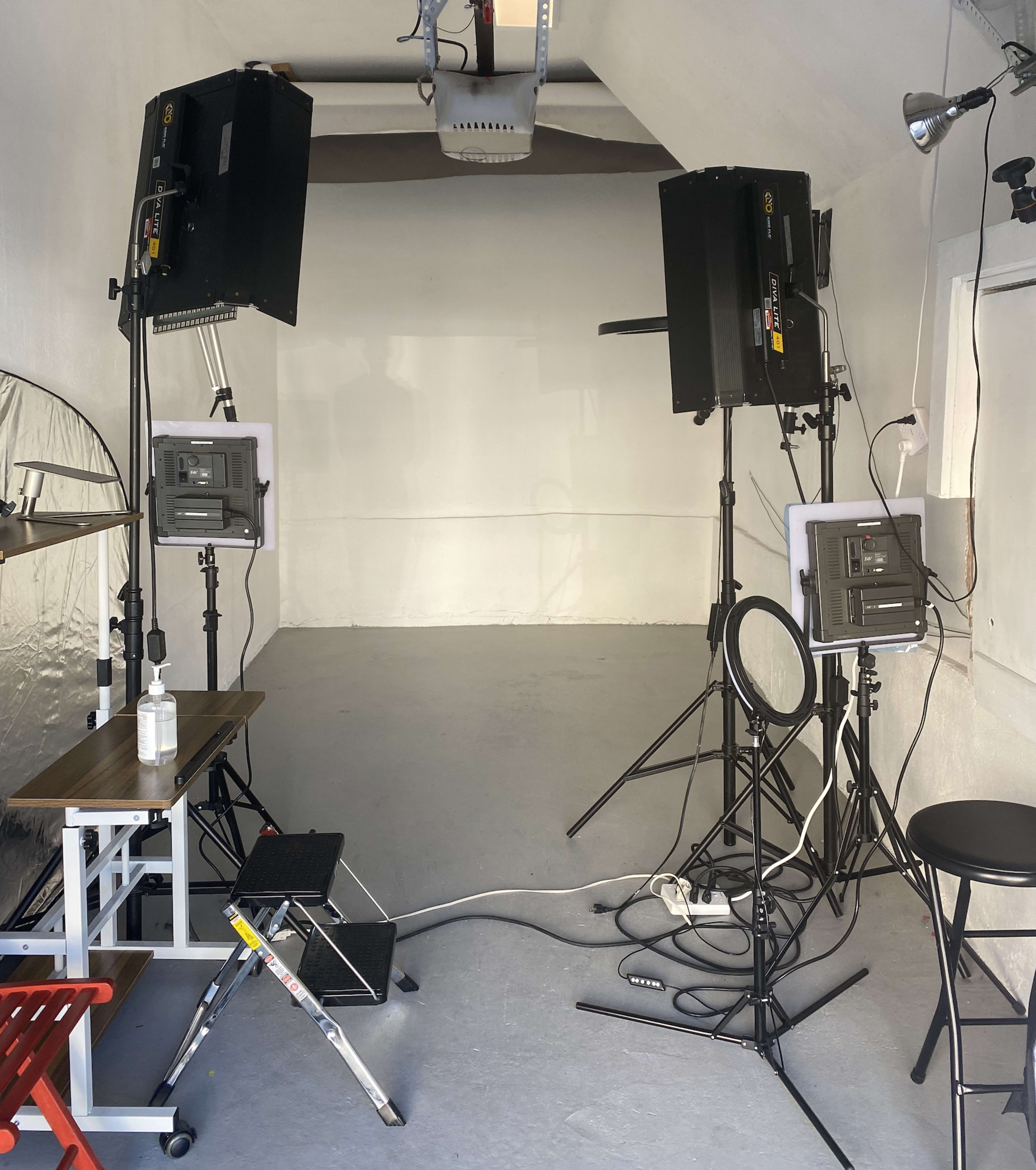 An empty, well-lit studio space equipped with two tall lights on stands, a ring light, and a small table with a stool and a ladder against the wall.