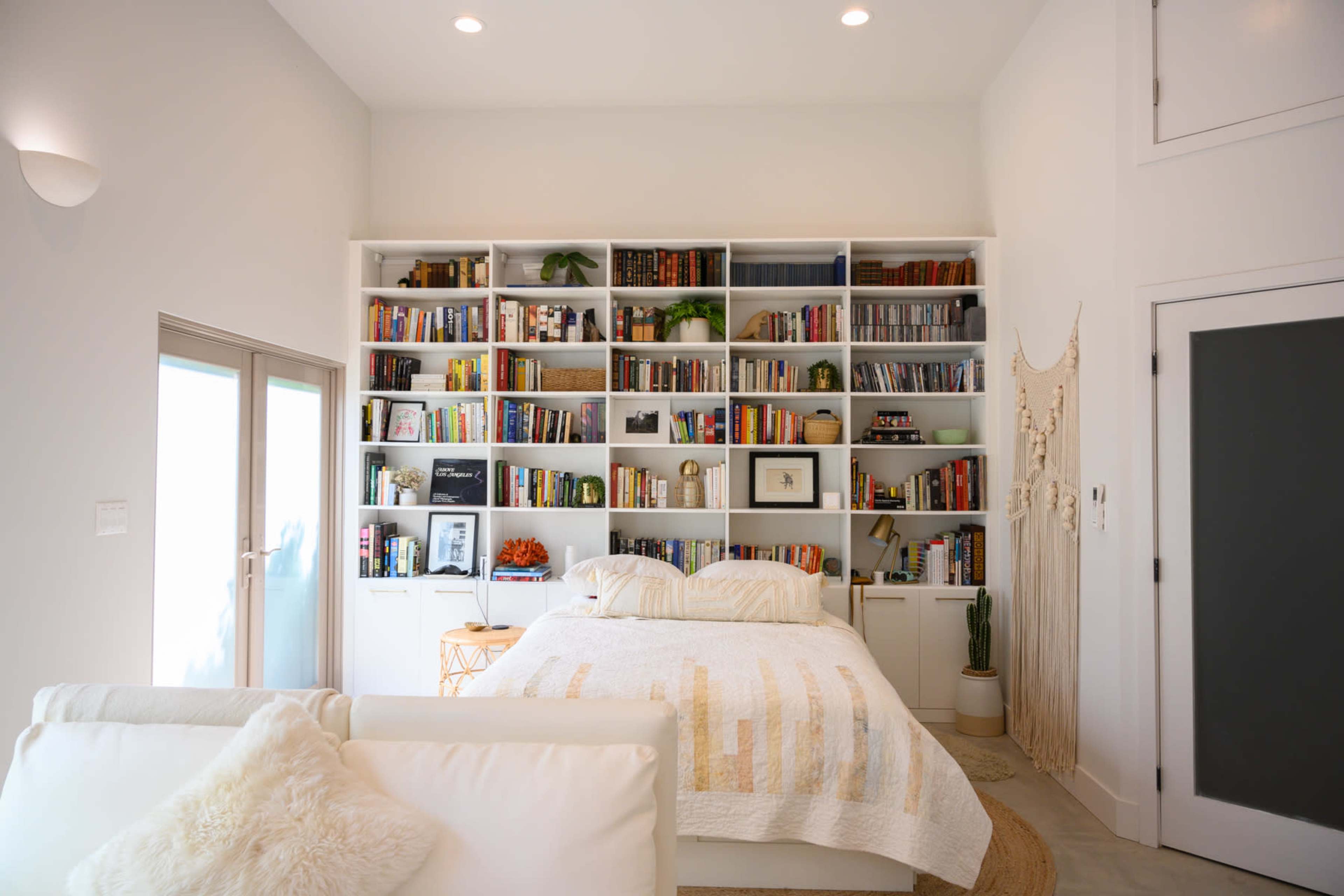 A neatly arranged bedroom features a bed in front of a large bookshelf filled with books and decorative items, alongside a cozy seating area.