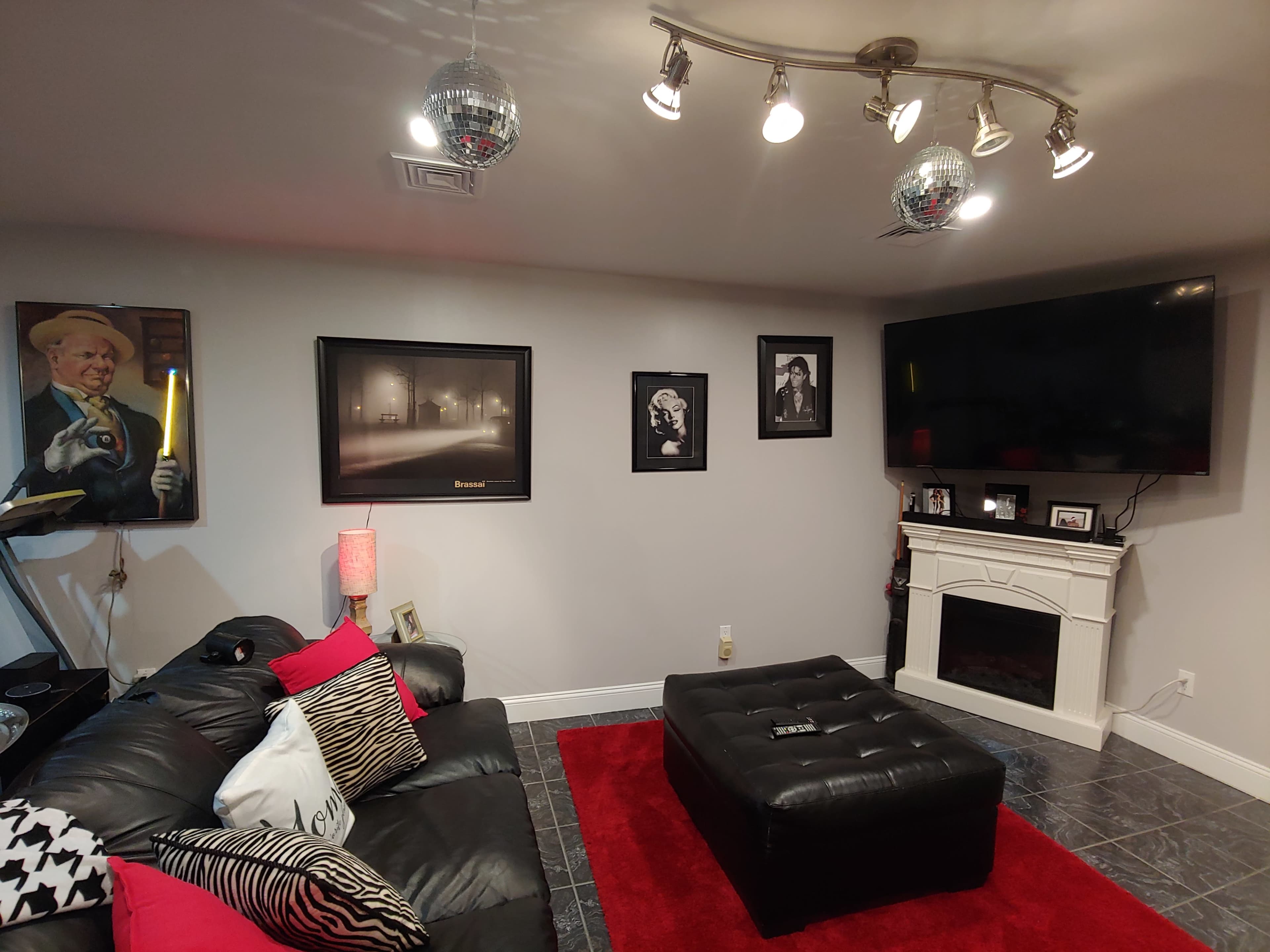 A modern living room with a black sectional sofa, a large television mounted on the wall, framed pictures, and a red area rug on a dark tiled floor.