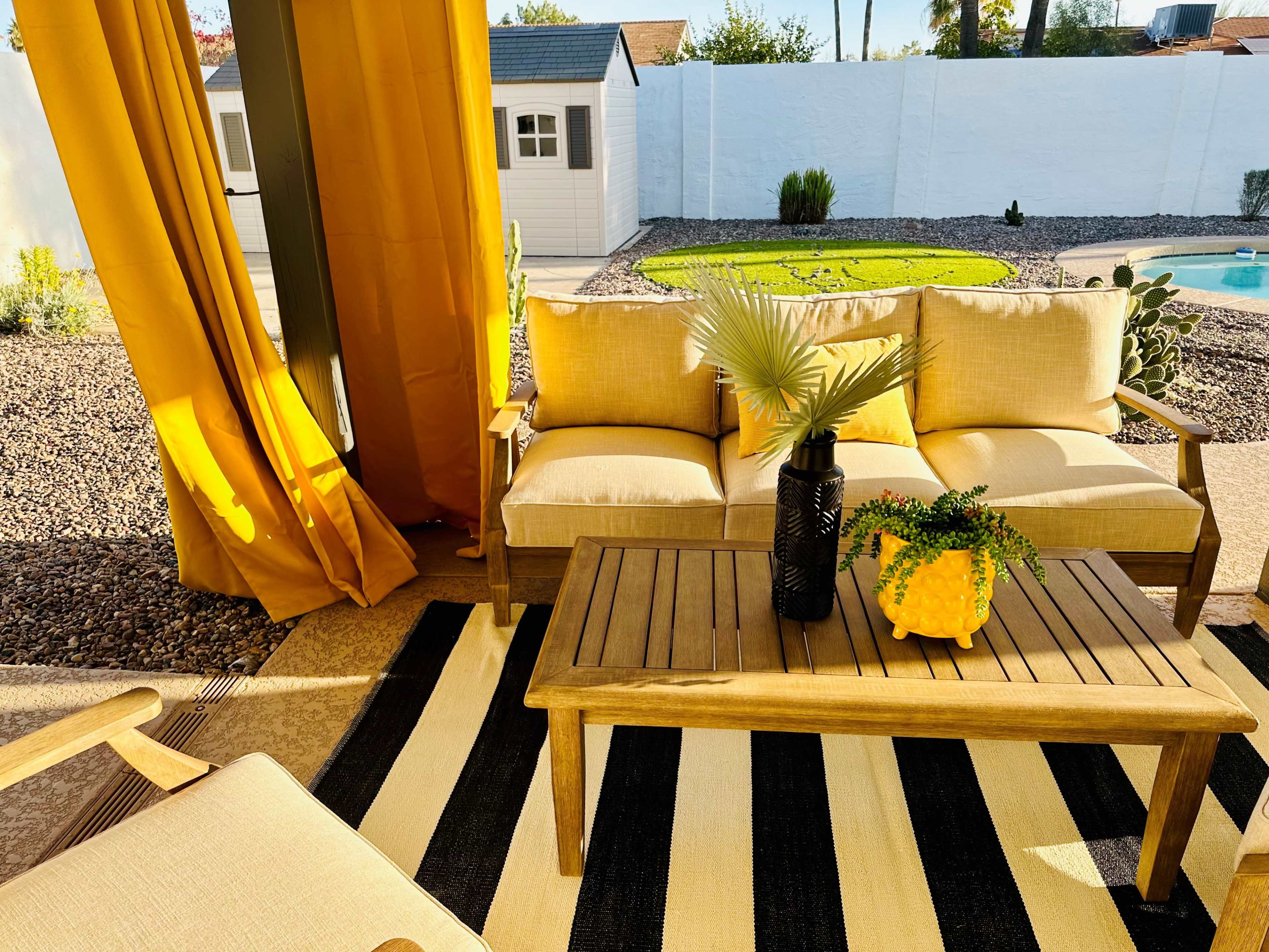 A patio seating area features a wooden table and couch with yellow cushions, surrounded by a striped rug and yellow curtains, overlooking a landscaped yard.