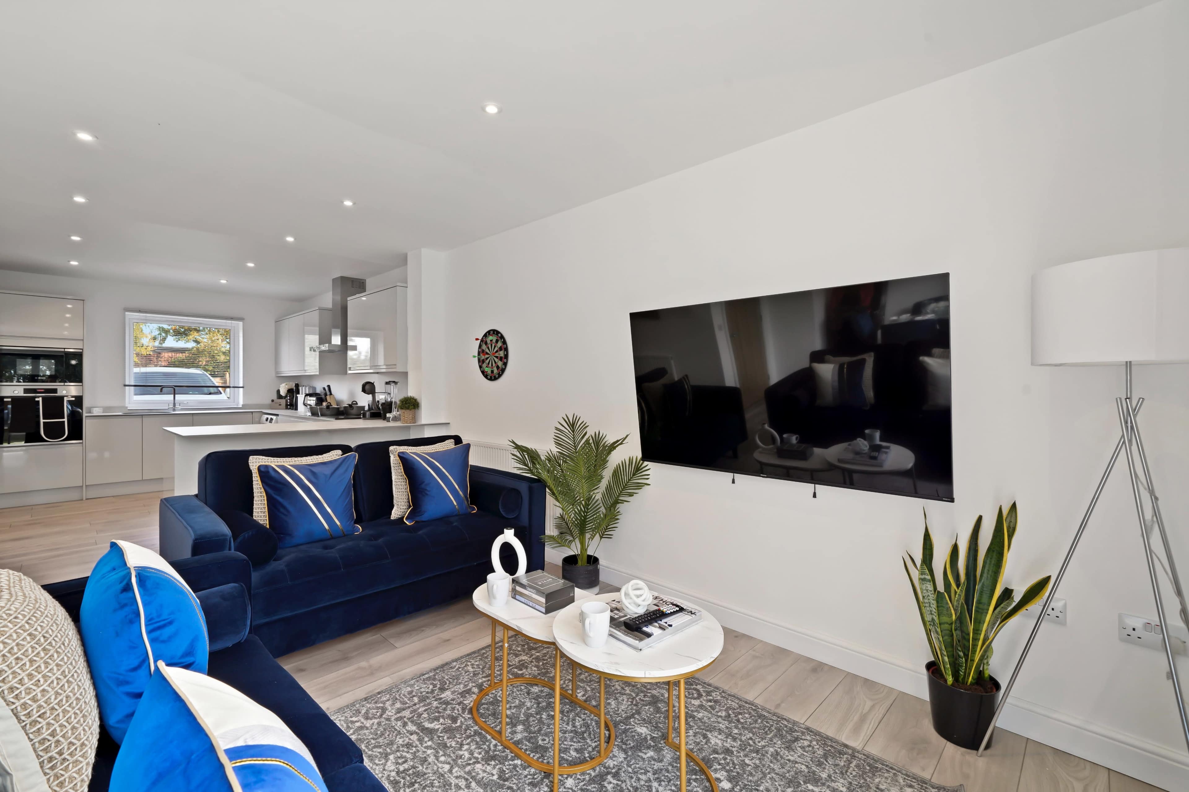 The image shows a modern living room featuring a blue sofa, a wall-mounted television, and a small coffee table with decorative items, adjacent to a well-lit kitchen area.
