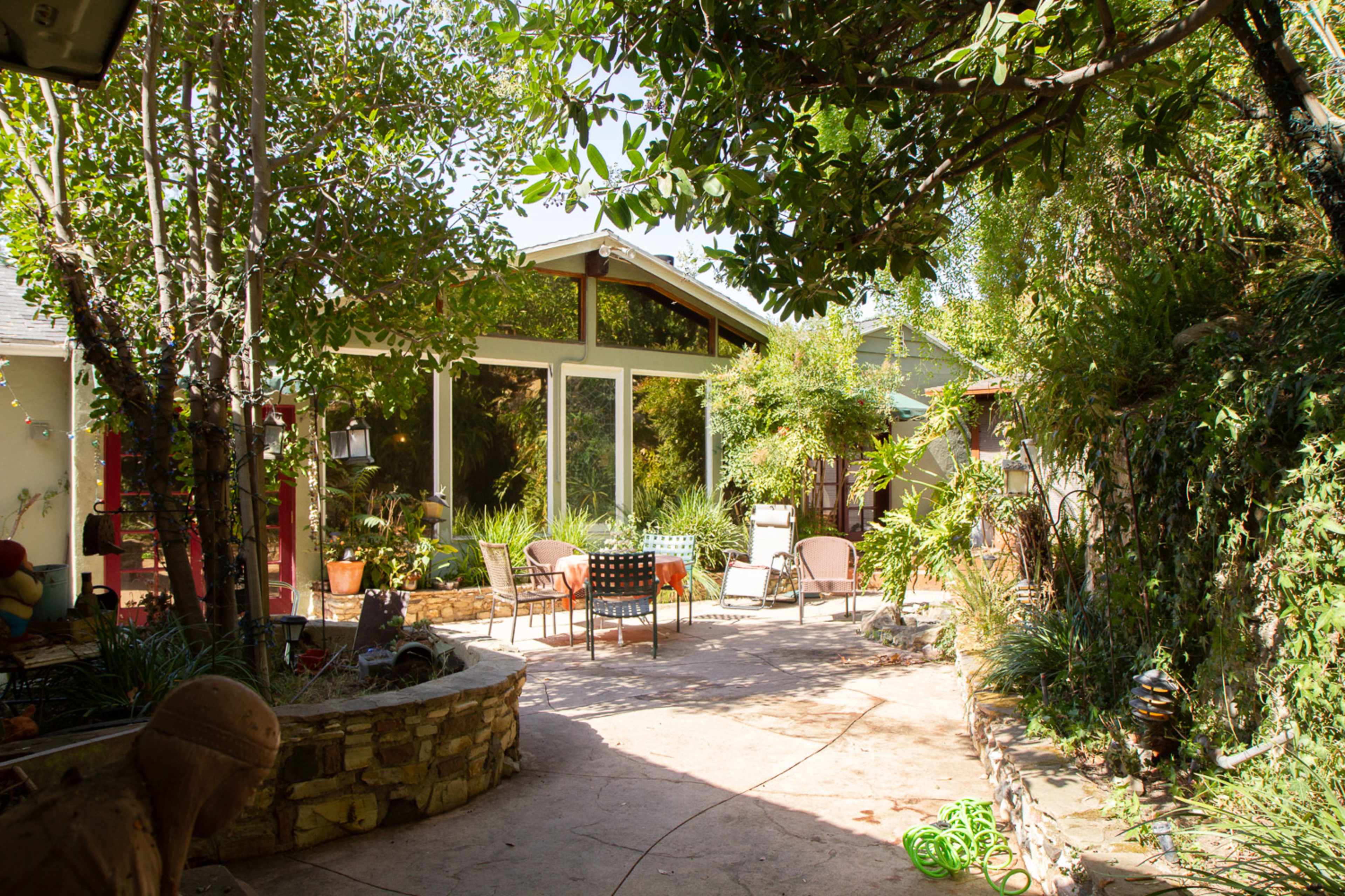 A patio surrounded by greenery features a seating area with chairs and a table, leading to a house with large windows.