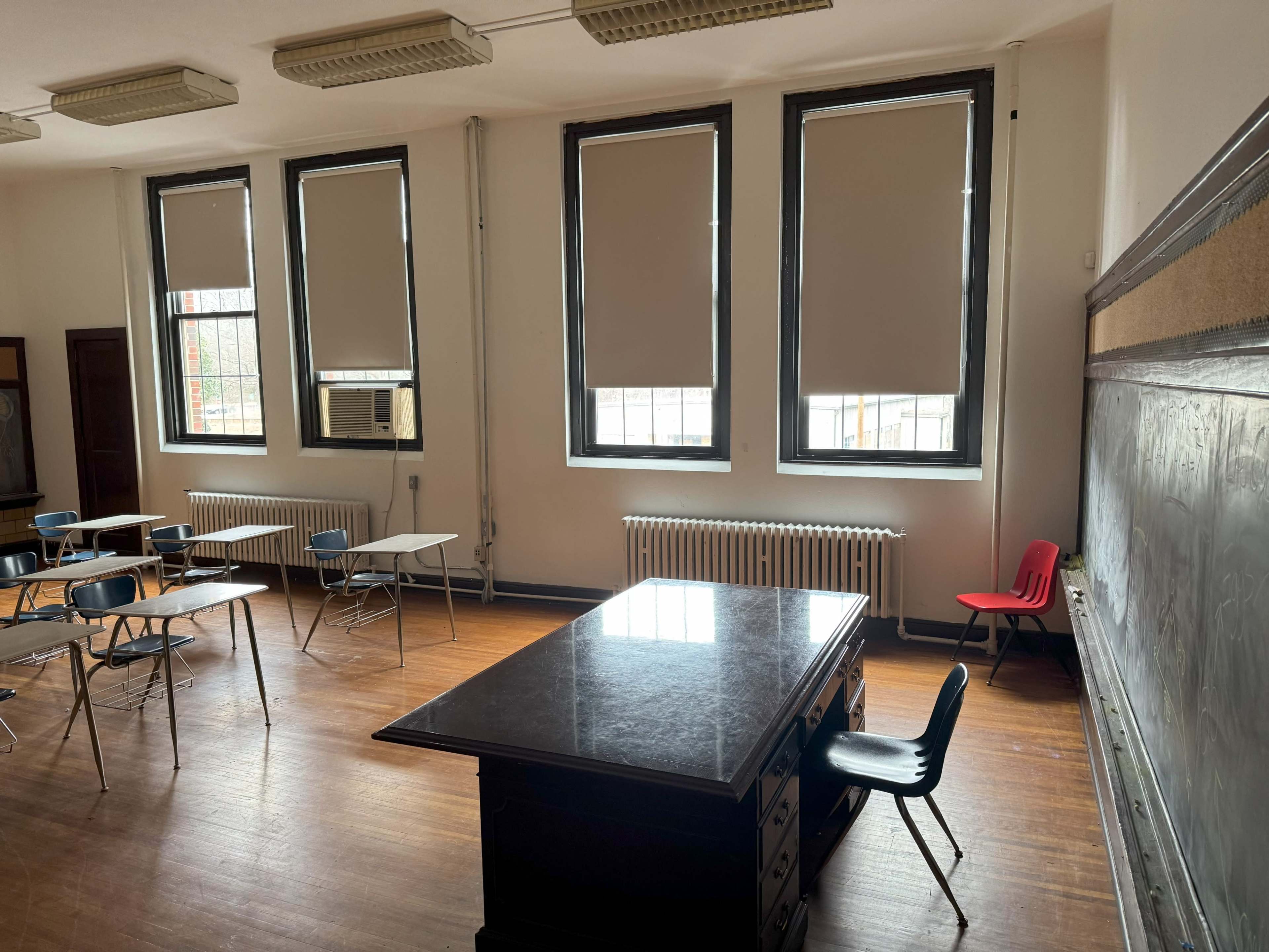 The image shows a sparsely furnished classroom with a large desk at the center, a few empty desks, and windows covered with closed blinds.