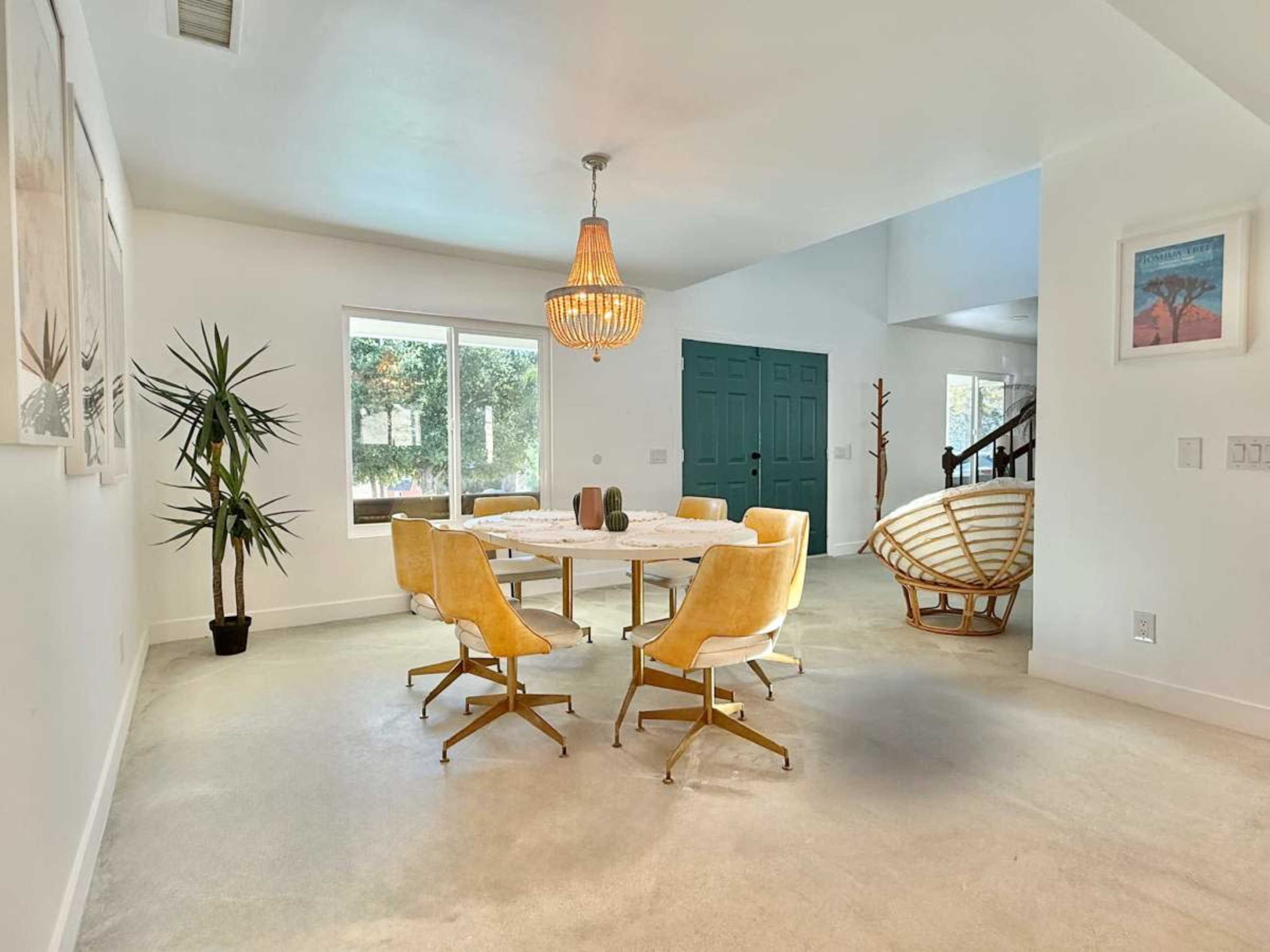 A modern dining area features a round table surrounded by yellow chairs, with a large window providing natural light and a view of greenery.