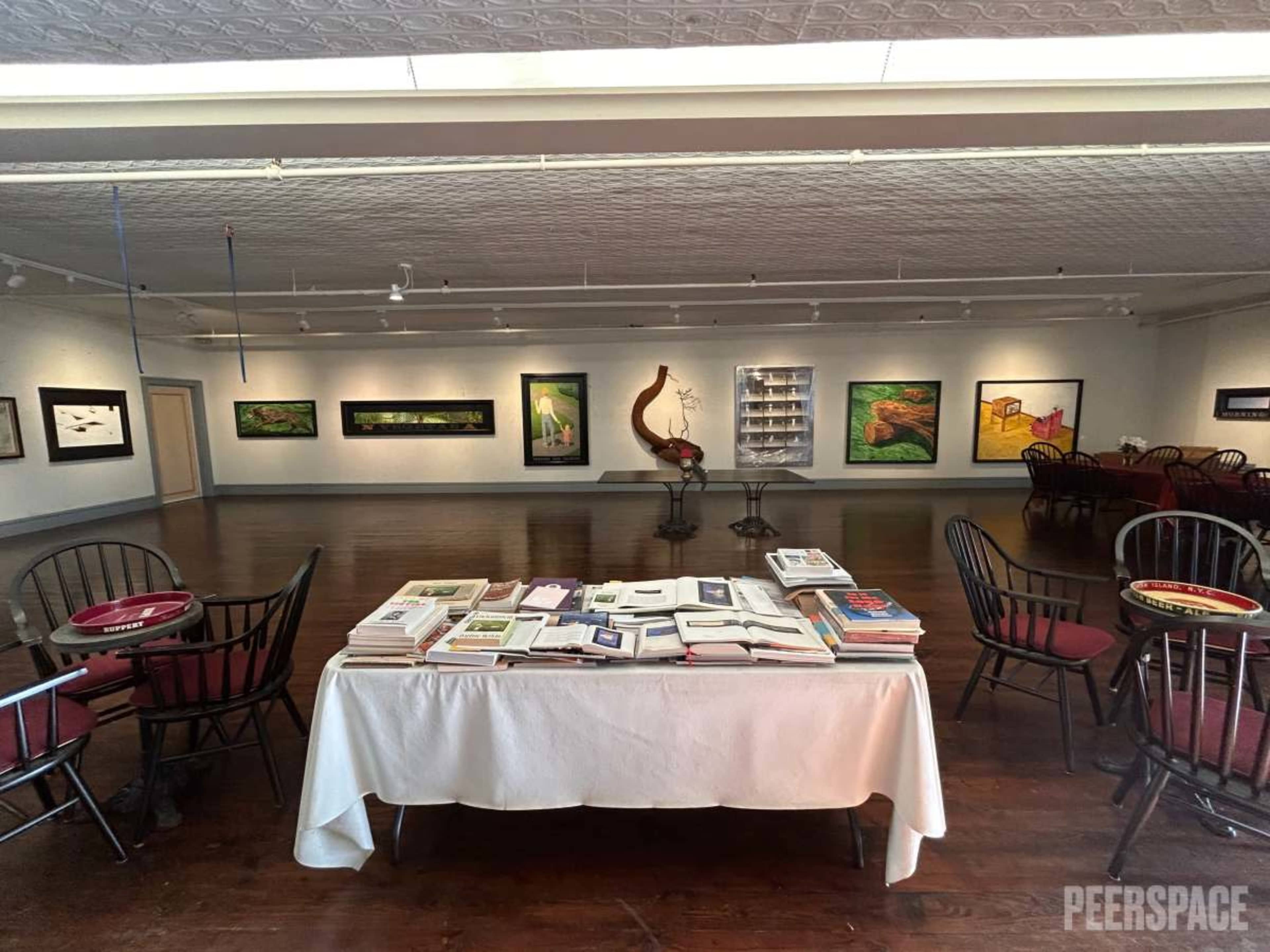 The image shows an art gallery with a table covered in books and brochures, surrounded by empty chairs and colorful paintings on the walls.