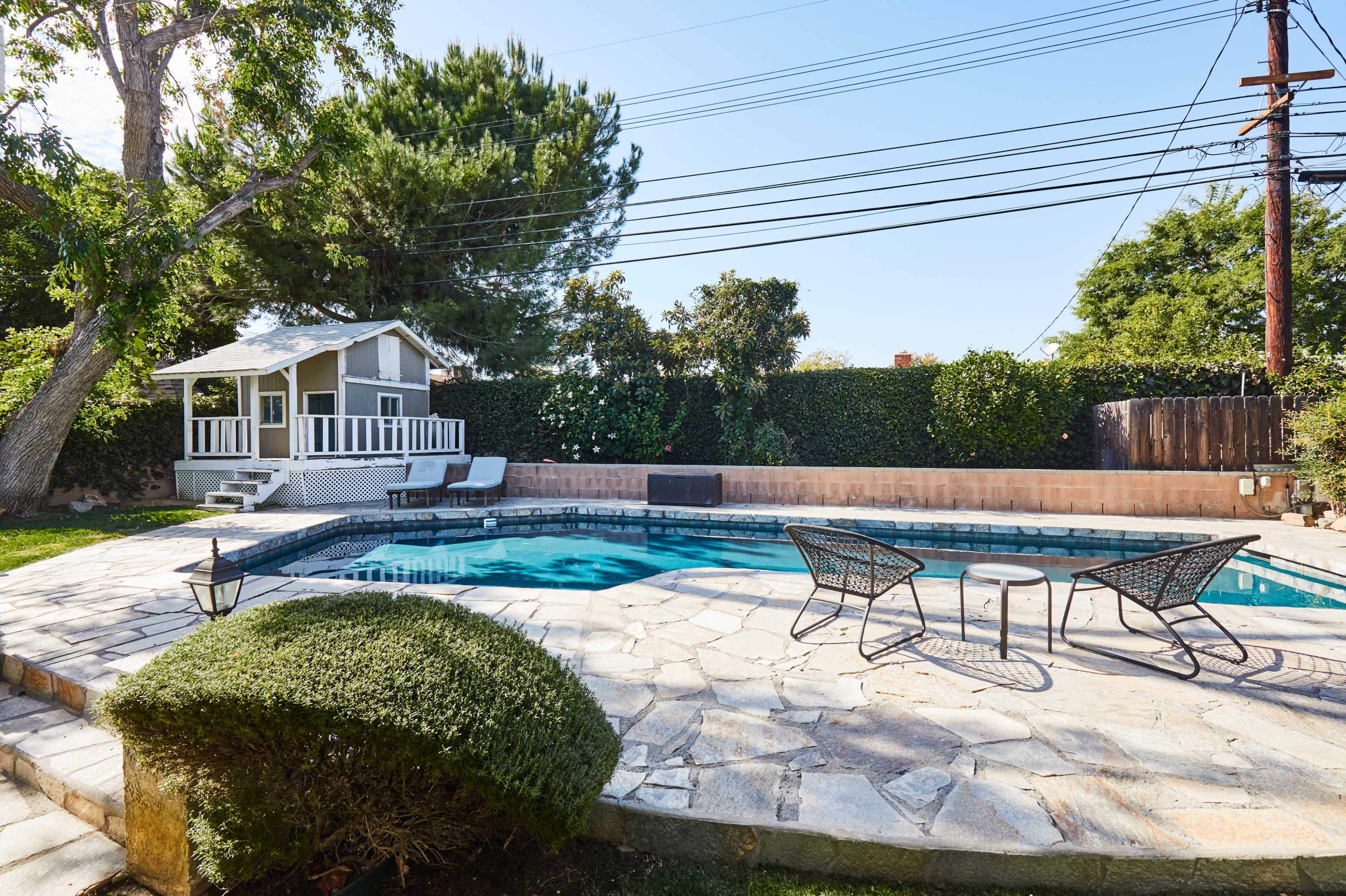 A landscaped backyard featuring a swimming pool surrounded by stone decking, with a small guest house and outdoor seating area nearby.