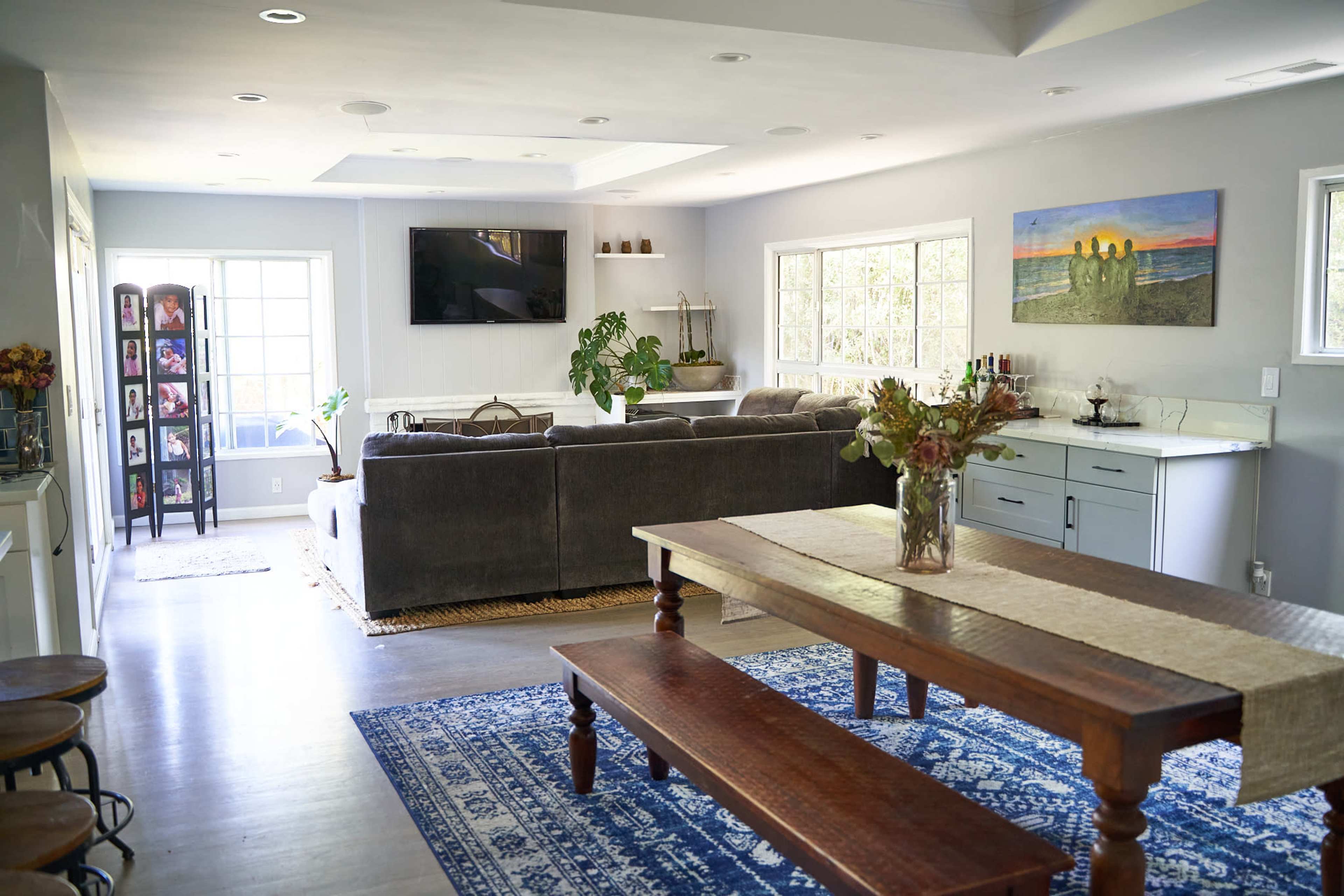 A modern open-concept living area features a gray sectional sofa, a wooden dining table, and a blue area rug, with large windows allowing natural light to fill the space.