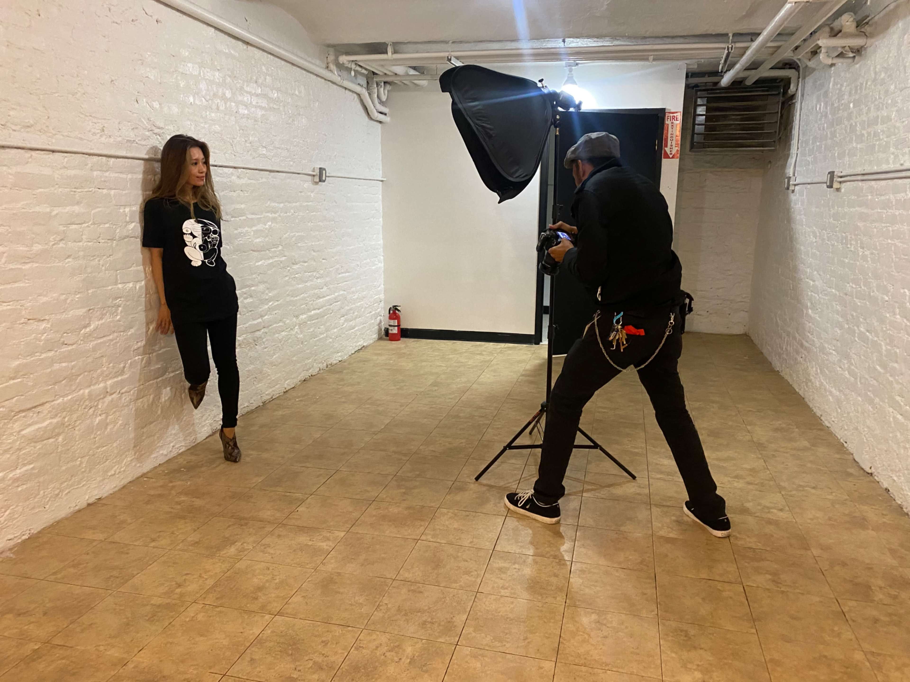 A photographer captures a model posing against a white brick wall in a sparse, well-lit studio space.