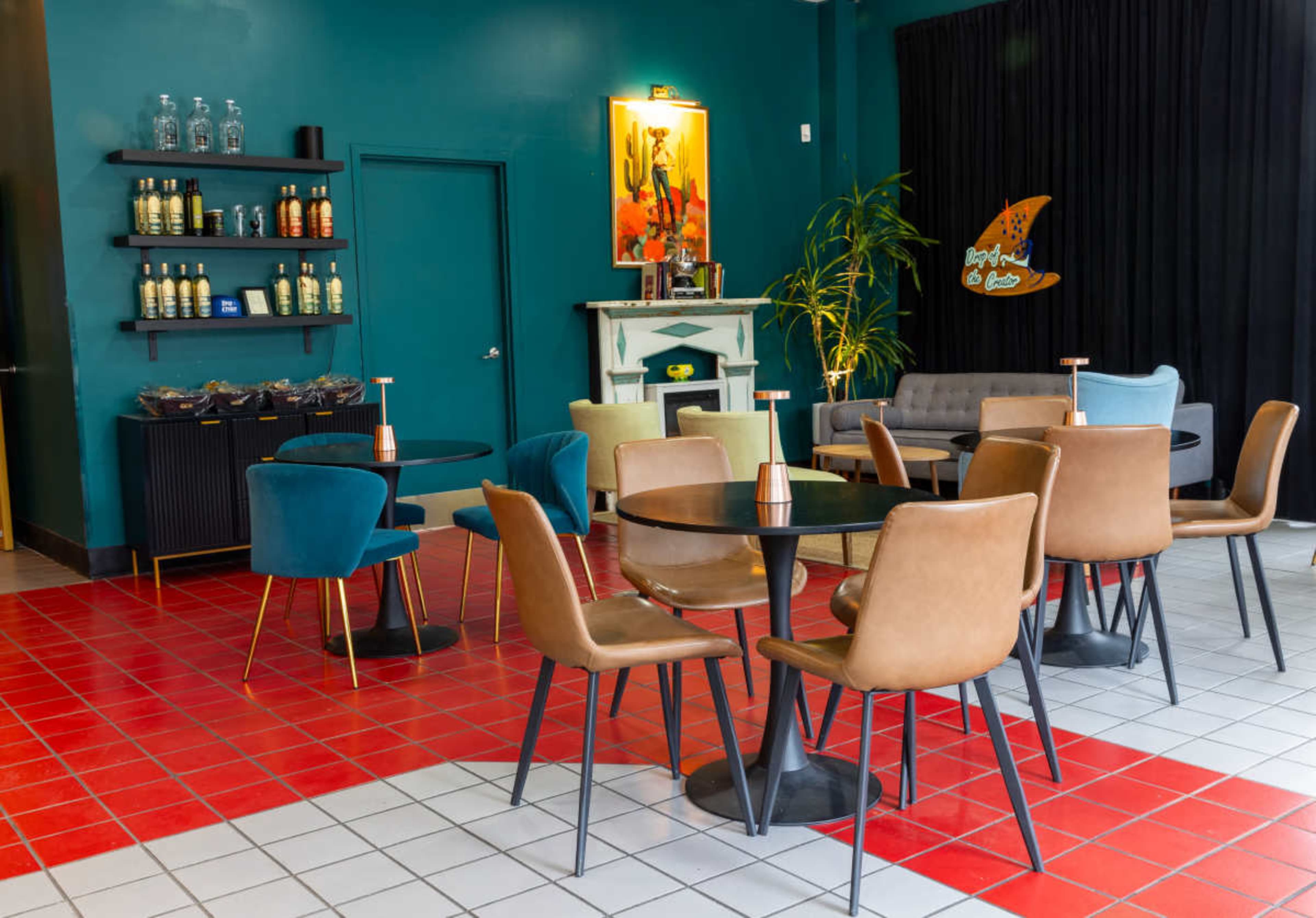 The image shows a brightly colored lounge area featuring several round tables with chairs, a bar shelf stocked with drinks, and a decorative wall with artwork.